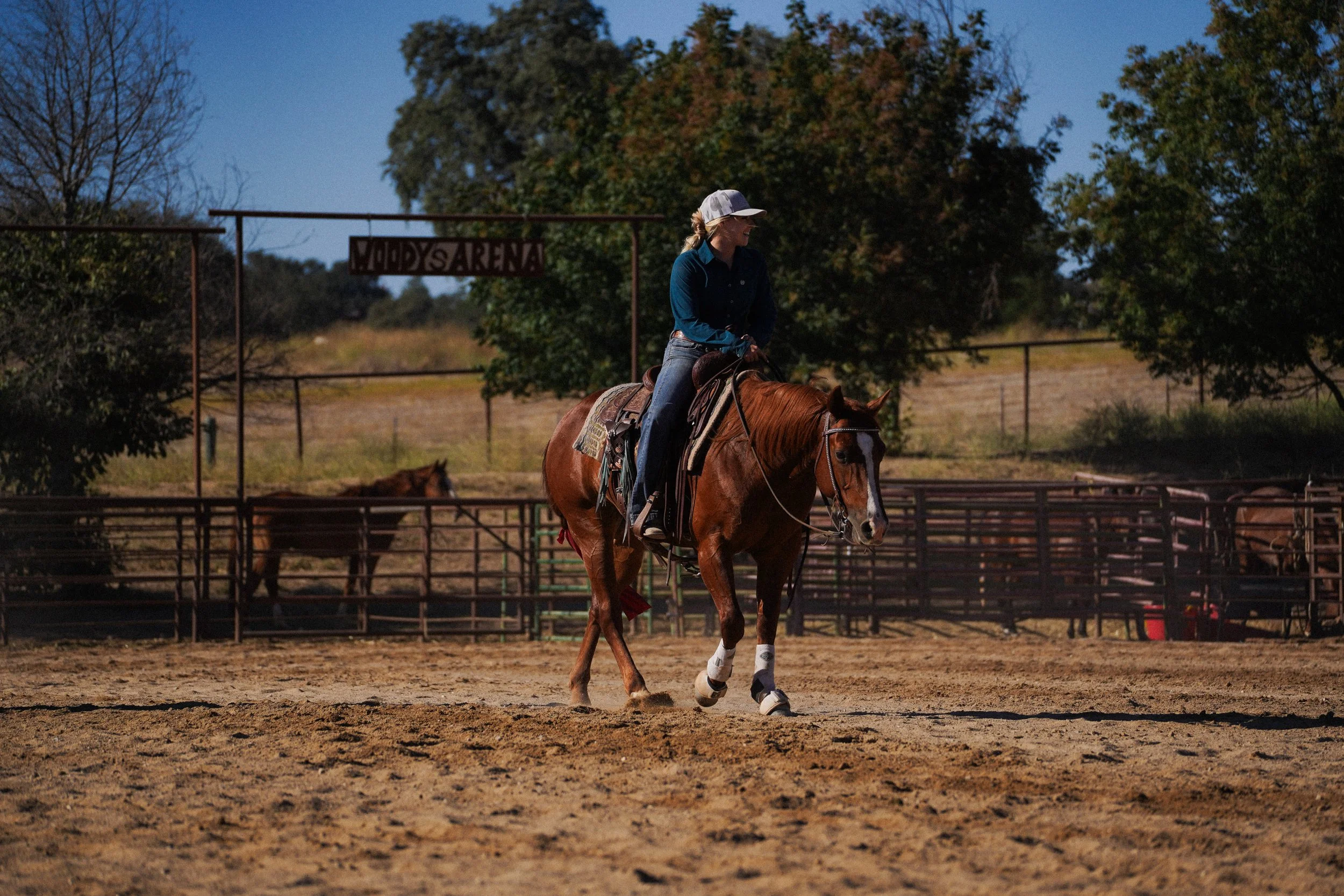 A woman riding a horse in an outdoor riding arena surrounded by trees and fencing.
