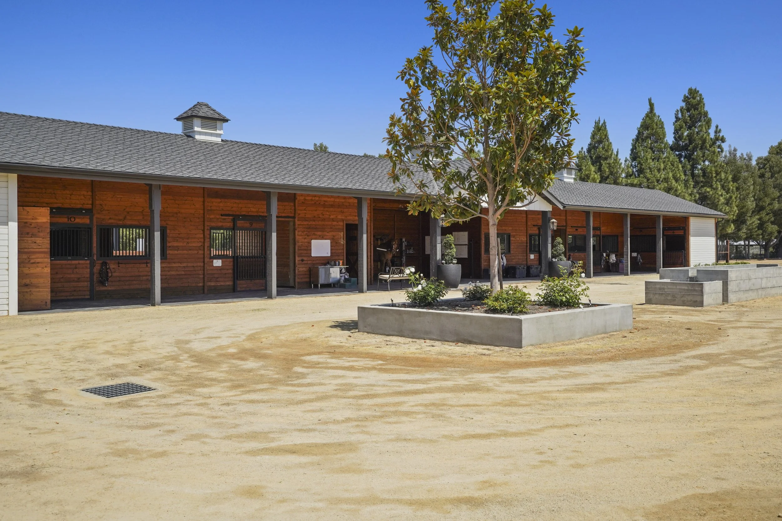 A stable with a wooden exterior and a gray shingled roof, surrounded by trees and concrete planters, under a clear blue sky.