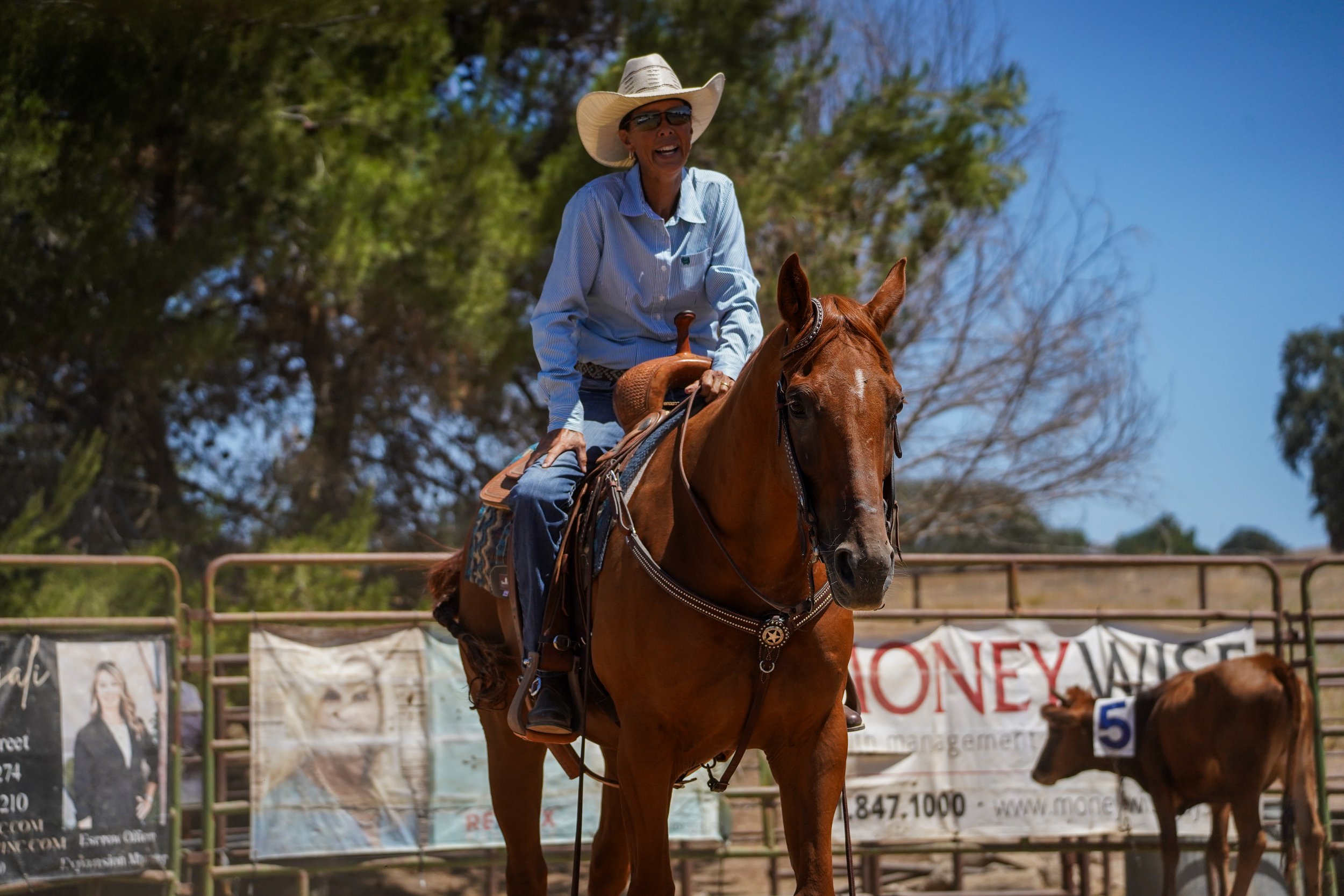A woman wearing a cowboy hat, sunglasses, and a light blue shirt rides a brown horse at an outdoor equestrian event. There are trees in the background and banners on the fence.