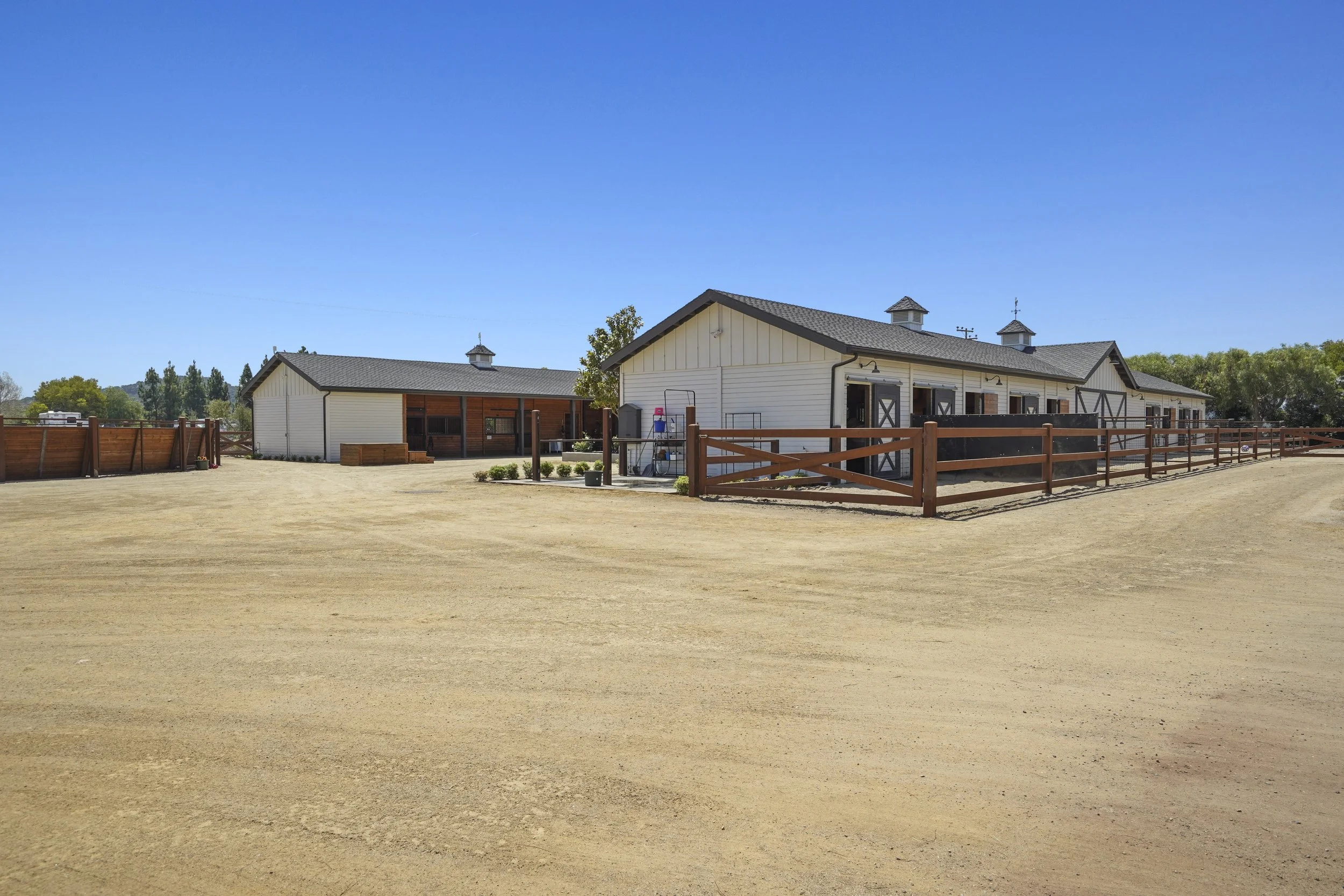 A large outdoor stable with white walls and dark shutters, surrounded by a fenced area, under a clear blue sky with some trees in the background.