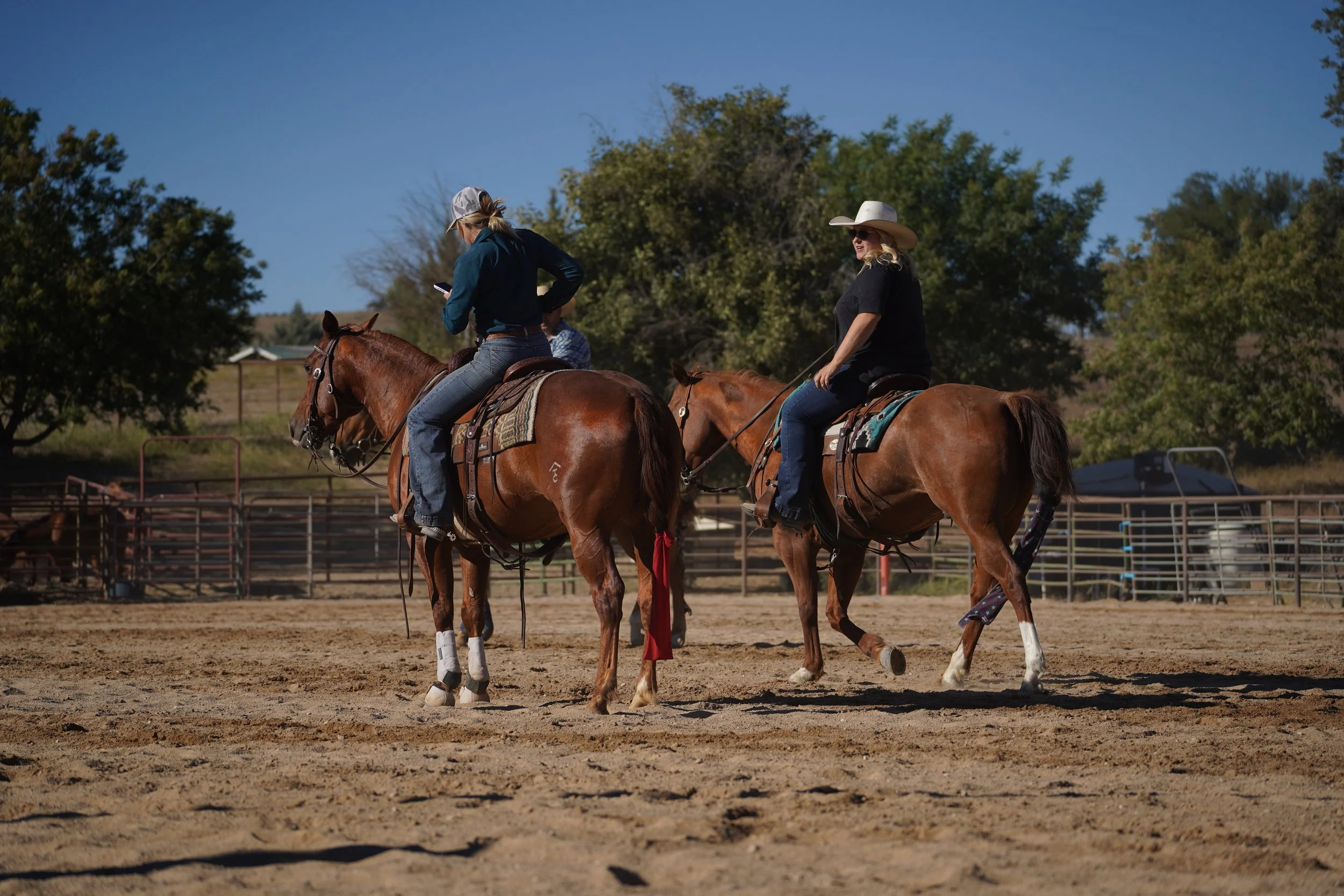 Two women riding horses in an outdoor riding arena with trees and a clear blue sky in the background.