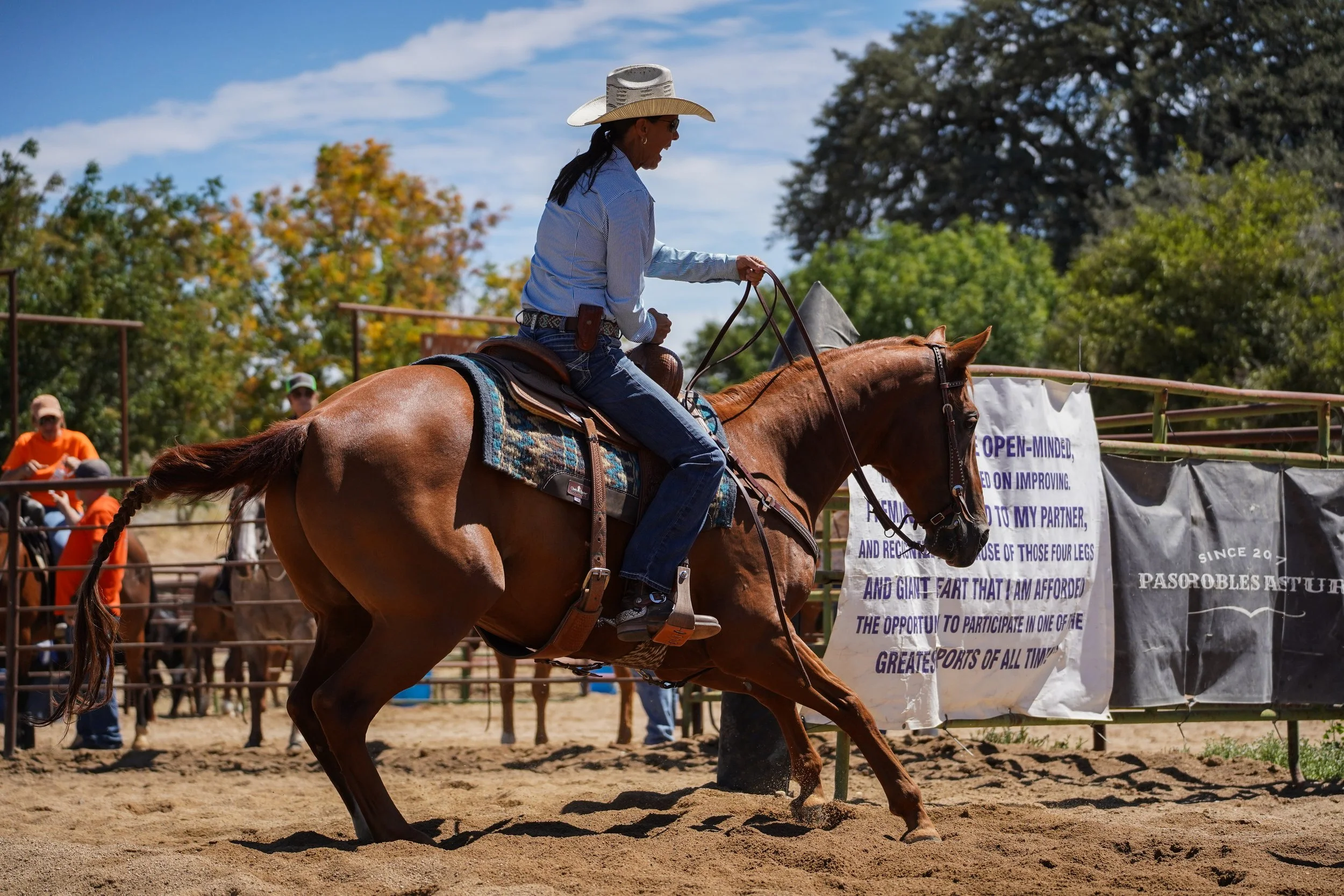 A woman riding a brown horse in an outdoor equestrian arena during daytime. She is wearing a wide-brimmed hat, sunglasses, a blue striped shirt, and jeans. There are spectators and banners in the background, with trees and blue sky above.