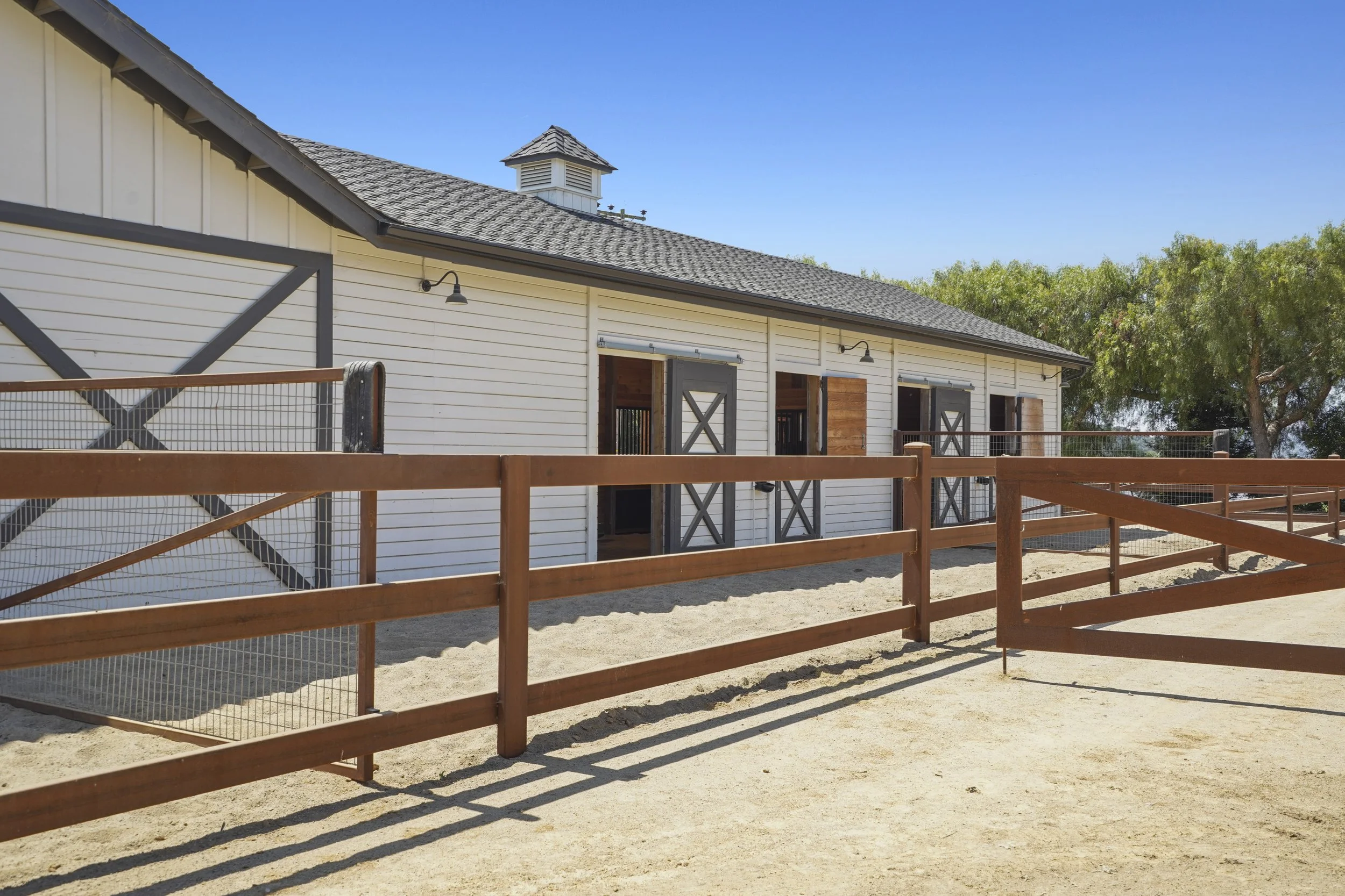 A white barn with gray trim and a gray shingled roof, surrounded by a wooden fence, with trees in the background and a clear blue sky above.