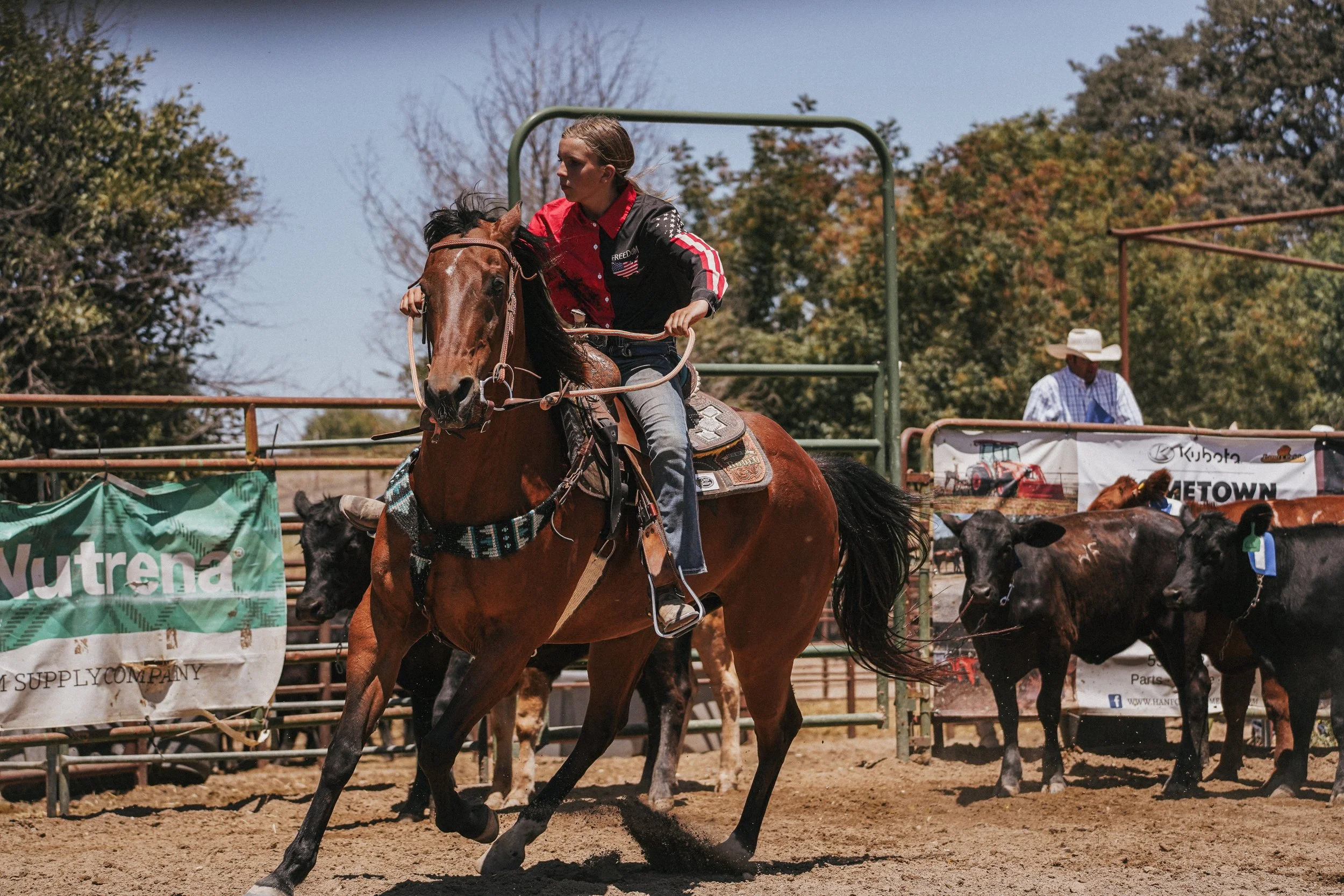 A young woman riding a brown horse in a rodeo arena with a cowboy in the background.