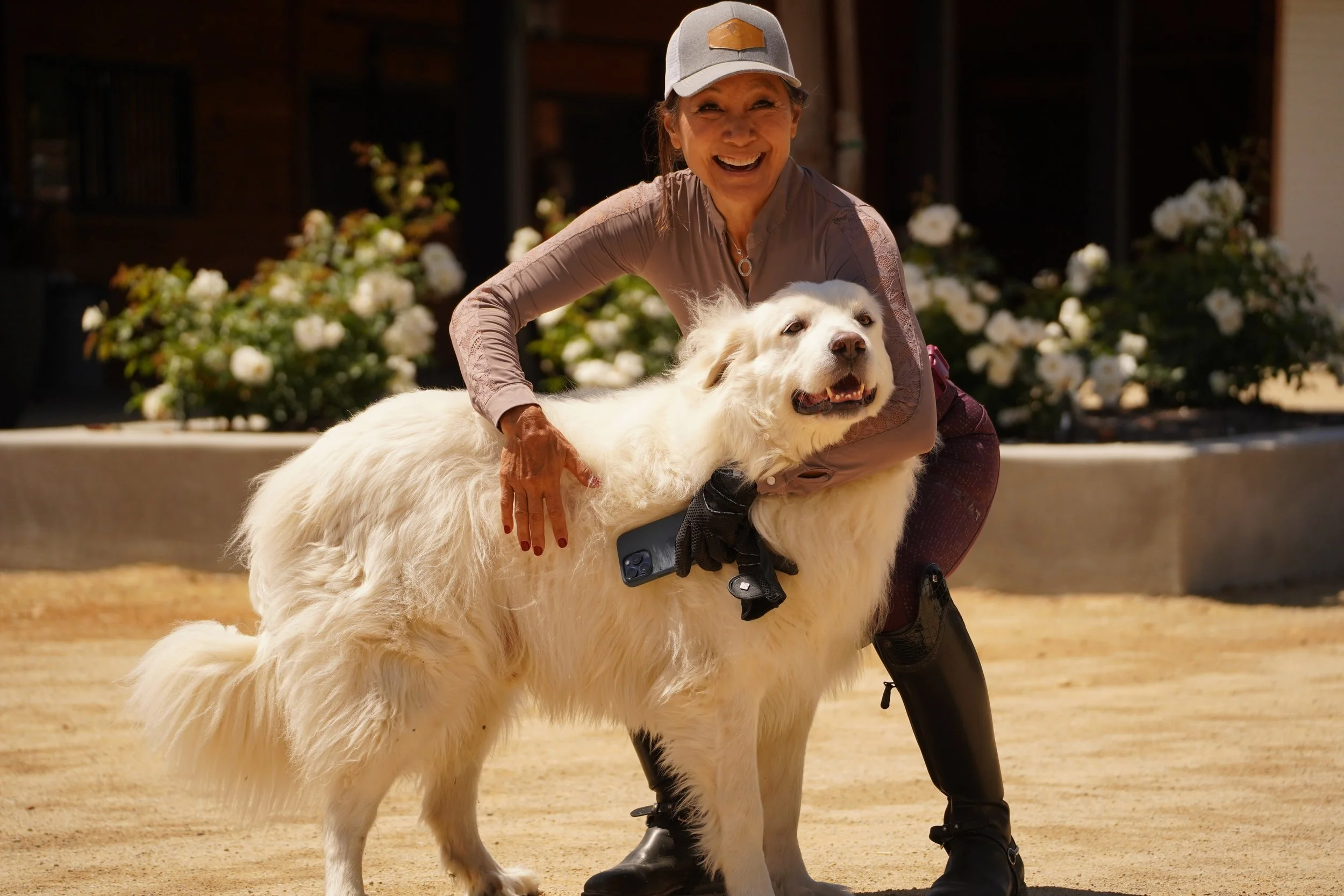 A woman in a baseball cap smiling and hugging a large white dog with fluffy fur outdoors, with white flowers in the background.