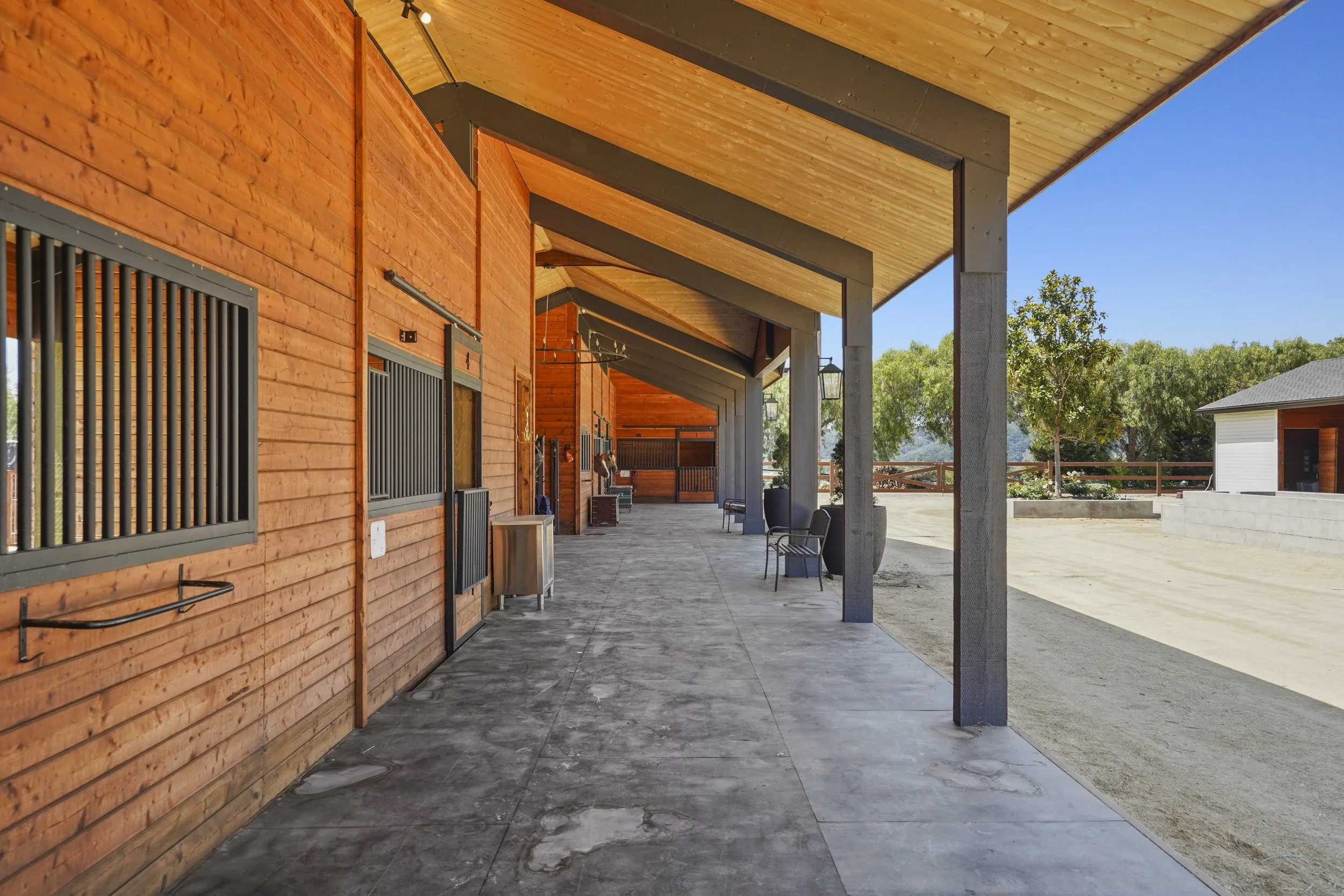 A covered porch area with wooden walls and ceiling, concrete flooring, and outdoor seating. Beyond the porch, there are trees and a small building with a shingled roof under a clear blue sky.