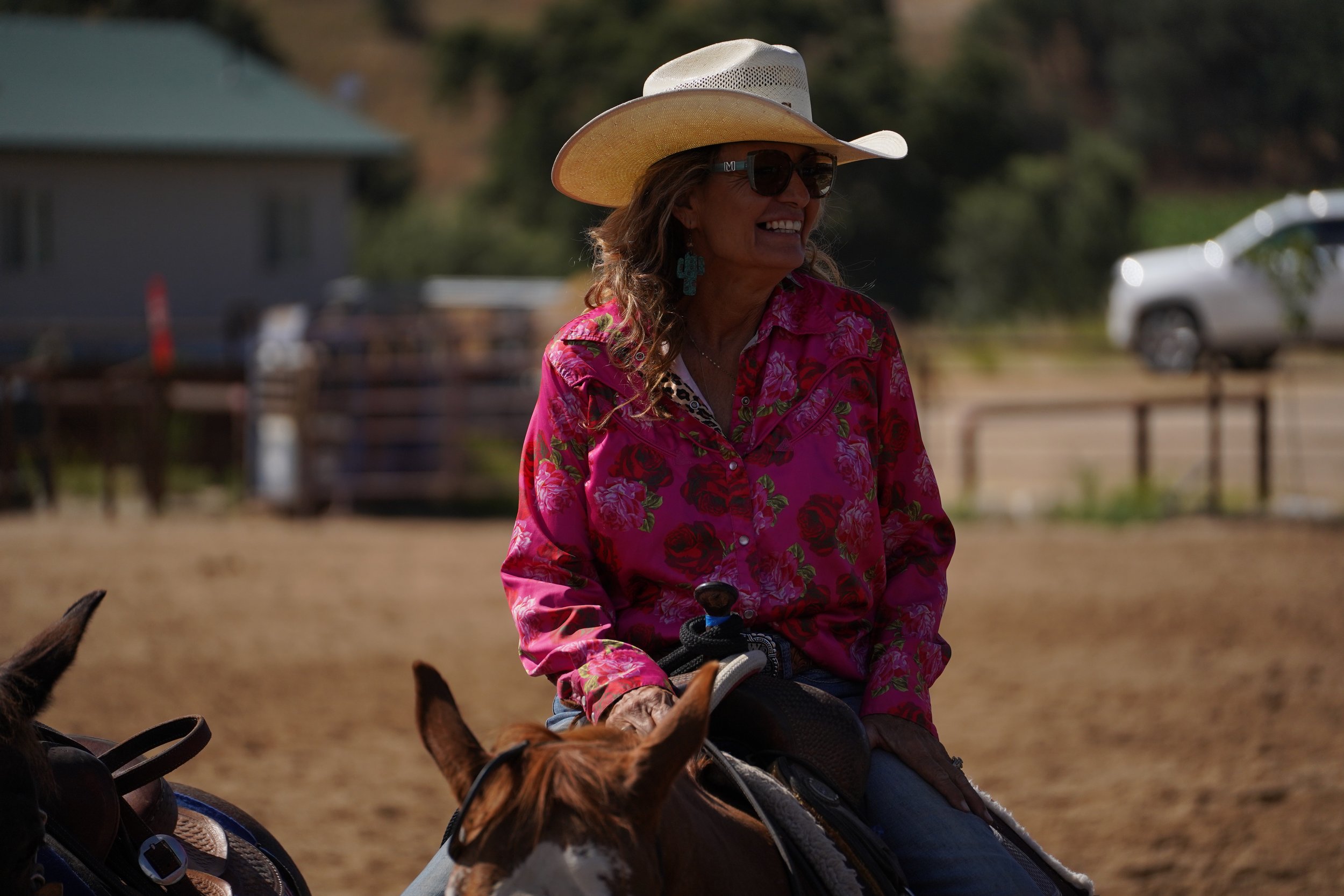 A woman wearing a cowboy hat, sunglasses, and a colorful floral shirt is smiling while riding a horse outdoors.