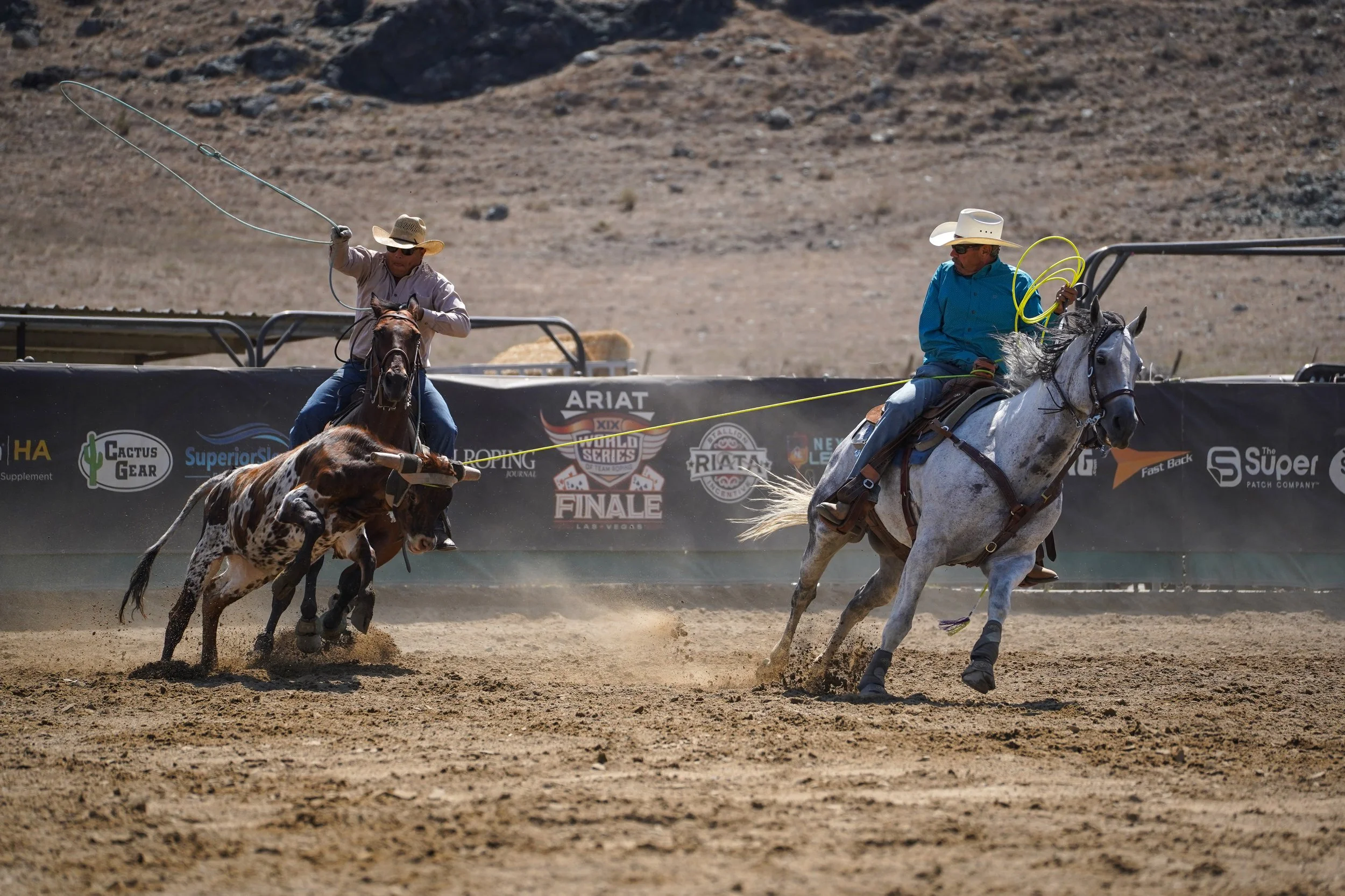 Two cowboys riding horses at a rodeo, one chasing a bucking bull on a dirt arena with a backdrop of mountains and banners.