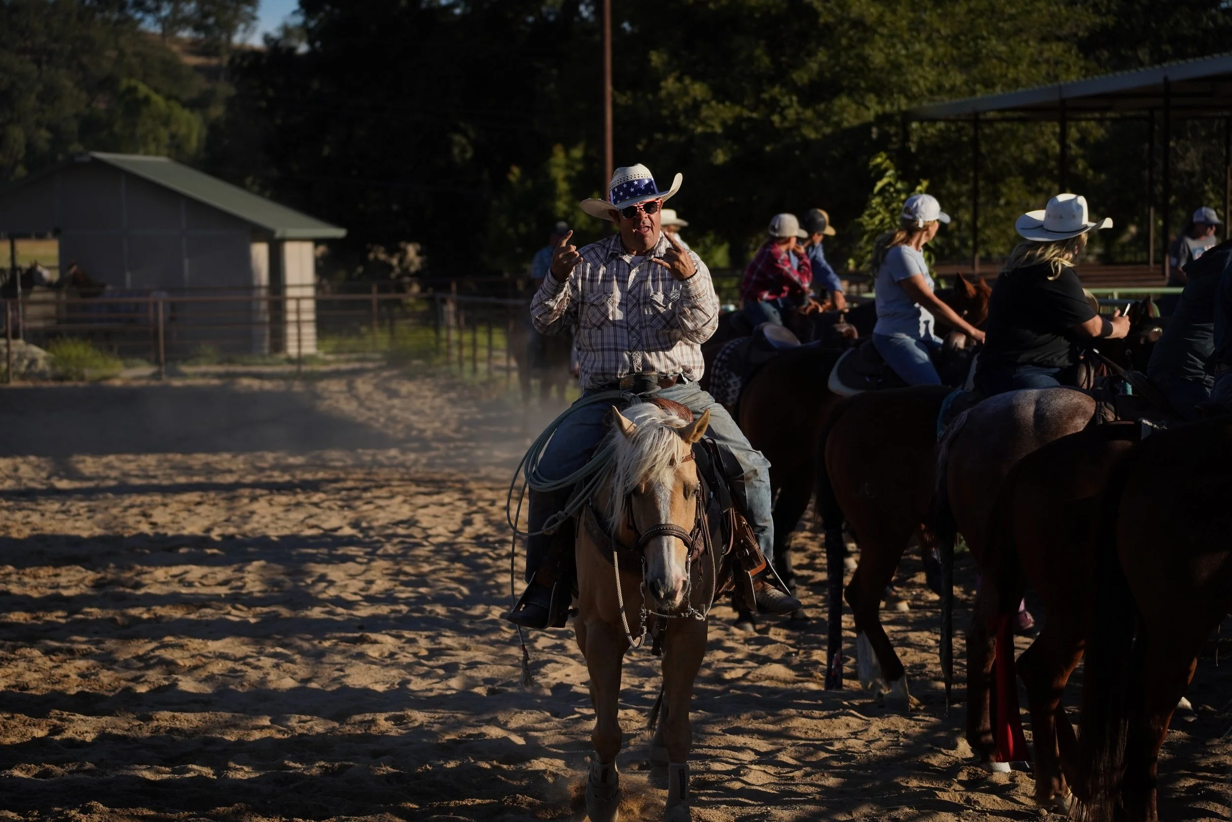 People participating in a horseback riding event outdoors. A man in the foreground is wearing a cowboy hat with American flag design, sunglasses, and a plaid shirt, riding a light-colored horse. Behind him, several riders are wearing cowboy hats, and
