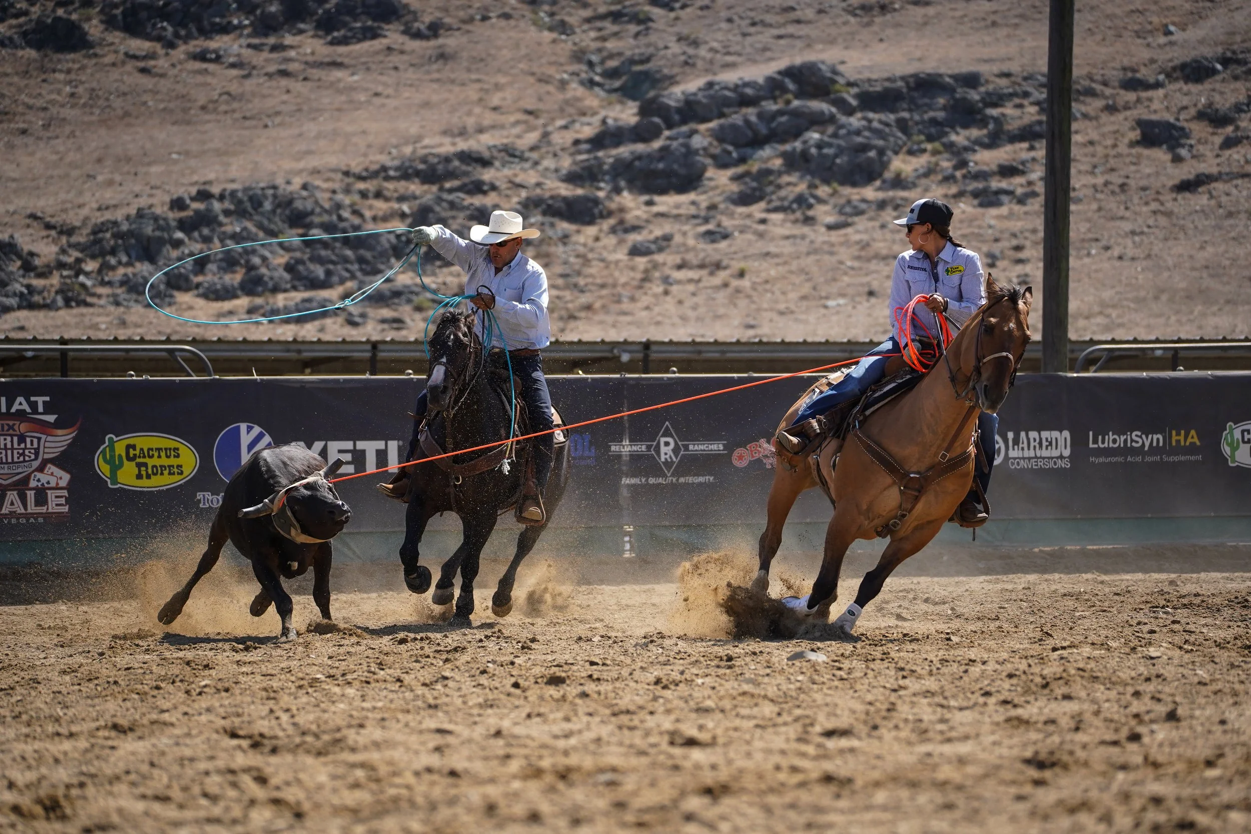 Two rodeo cowboys on horseback participating in steer roping on dirt ground with a background of rocky, arid landscape.