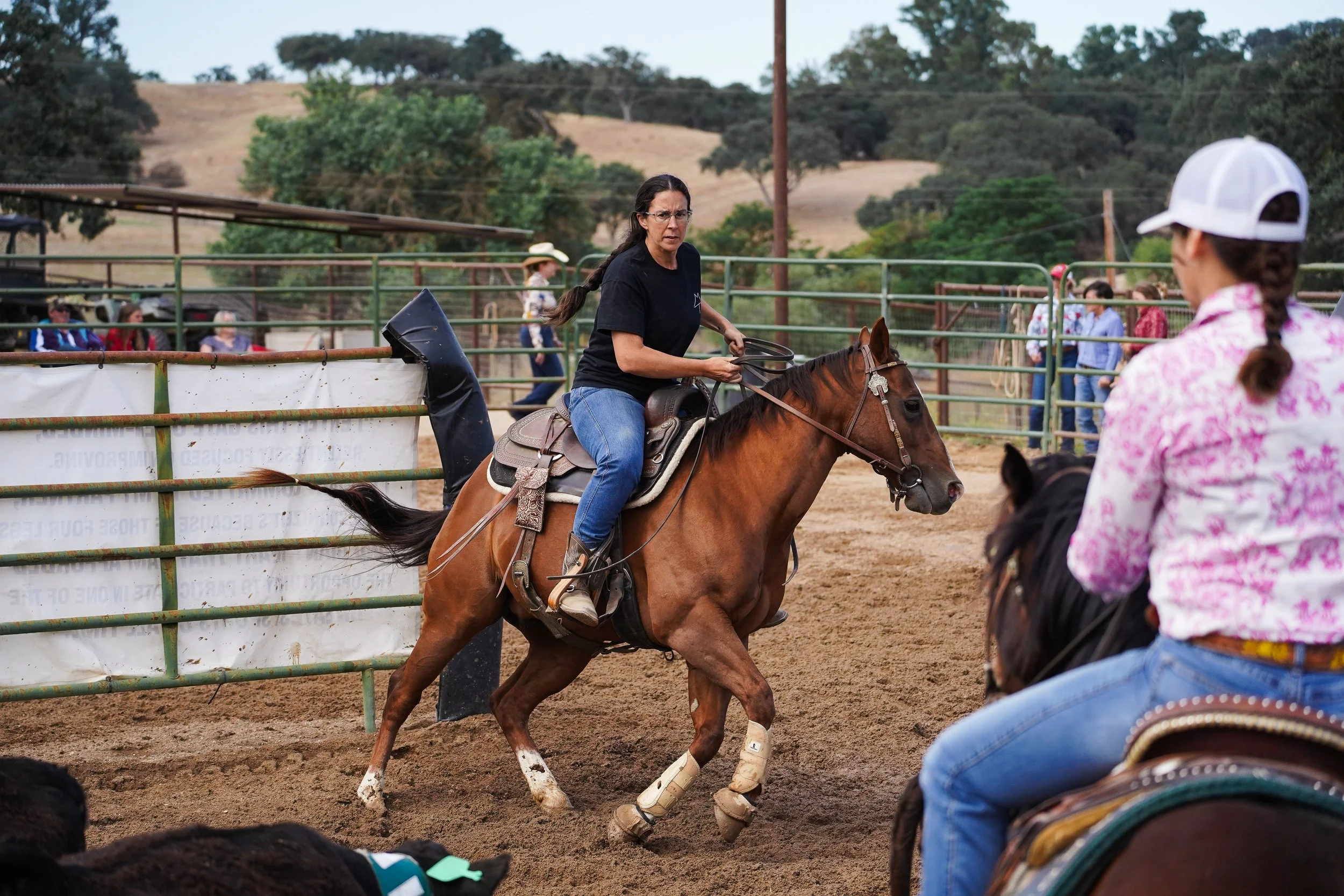 A woman riding a horse at a rodeo or equestrian event, with onlookers and fencing in the background, set outdoors with rolling hills and trees.