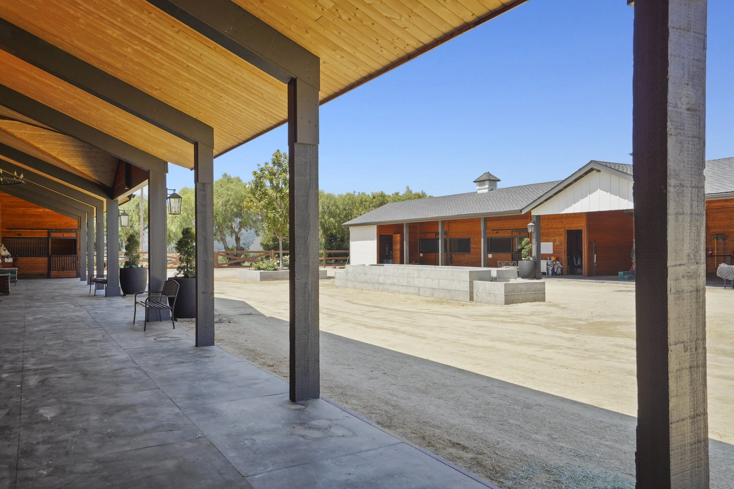 View of a construction site with a covered patio area, wooden beams, and a building with white walls and gray roof in the background, surrounded by trees and clear blue sky.