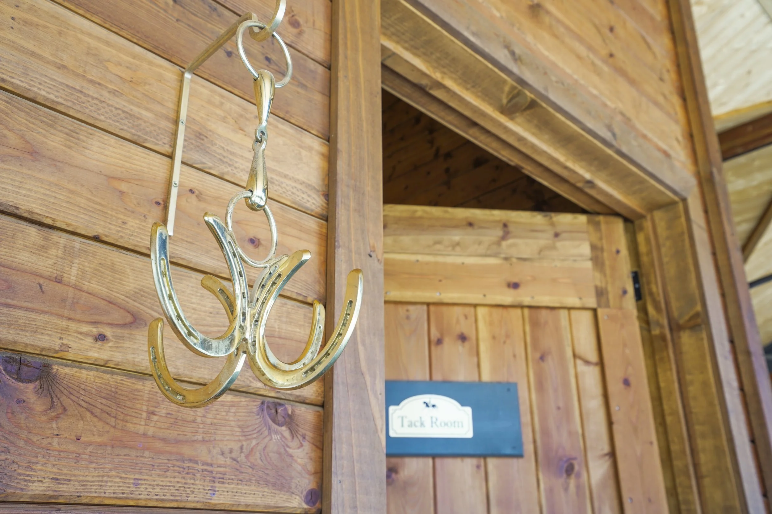 A metal coat hook shaped like a floral pattern is mounted on a wooden wall inside a rustic room or cabin. There is a sign indicating it is the Tack Room.