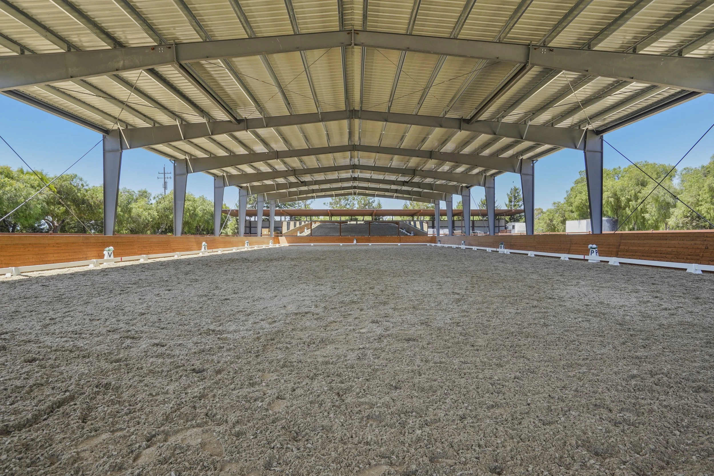 An indoor equestrian riding arena with a metal roof, wooden barriers, and a sandy ground surface, surrounded by green trees.