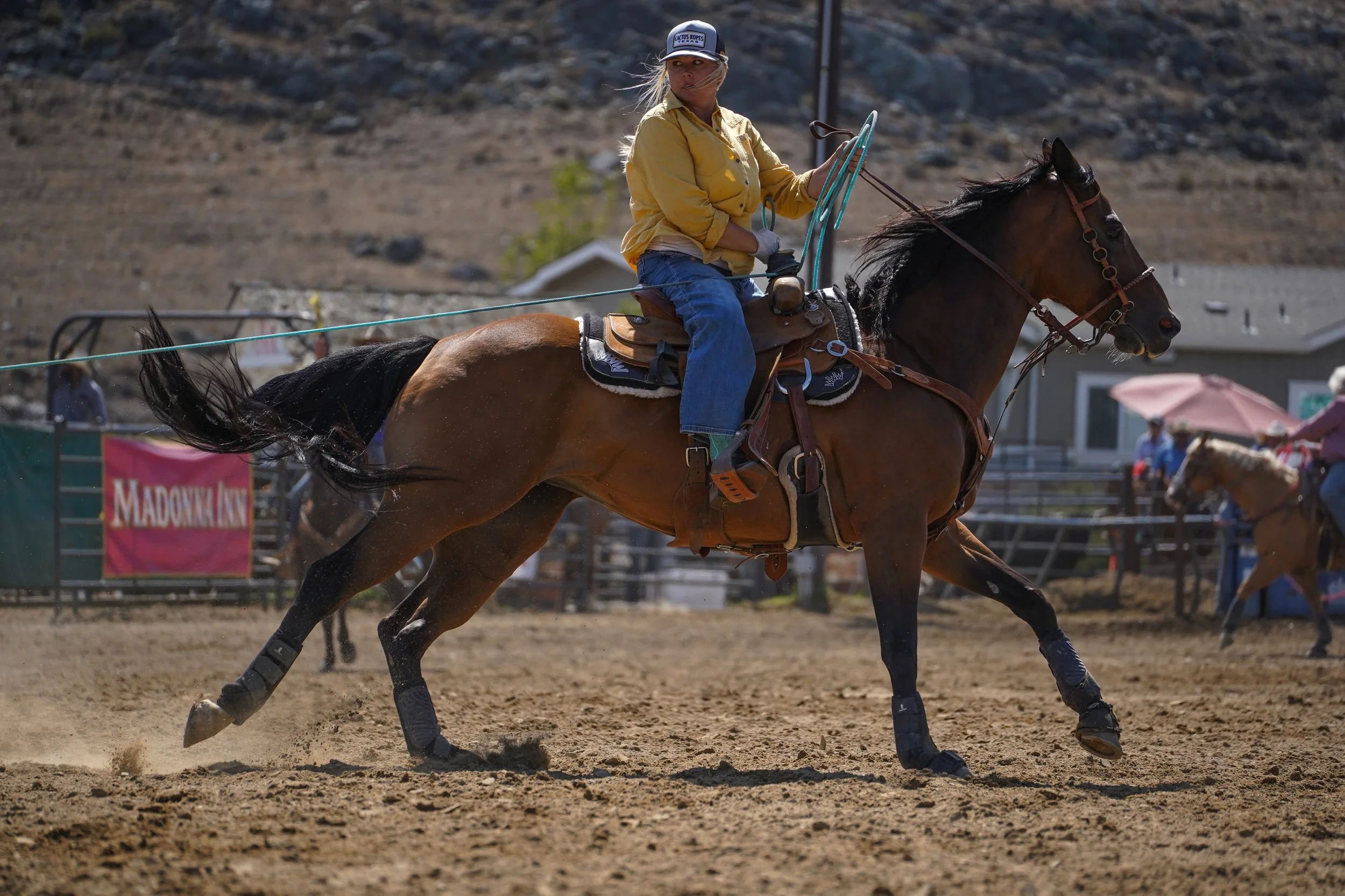 A woman riding a horse during a rodeo event on a dirt arena with a mountain background, wearing a cap, yellow shirt, and jeans.