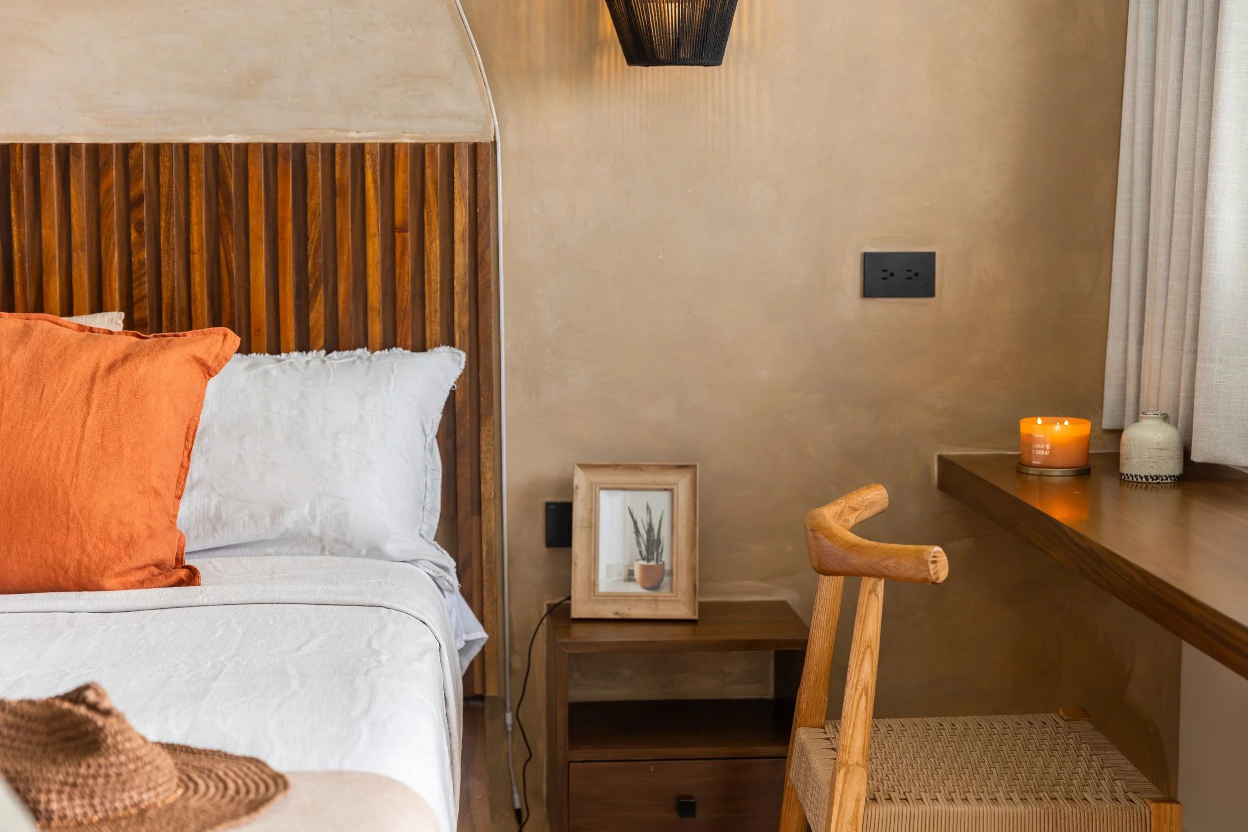 Real estate photography of a bedroom corner with a bed having white and orange pillows, a wooden headboard, a framed picture of a plant, a bedside table, a lit candle, a ceramic vase, a wooden chair, at Mahahual.
