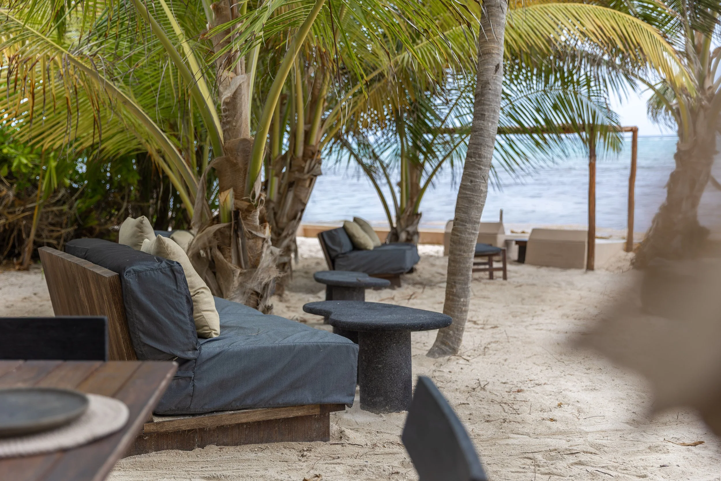 A beachside outdoor seating area with black cushioned sofas, pillows, and small round tables under palm trees, overlooking the ocean with a wooden frame in the background, at Bespoke Tulum.