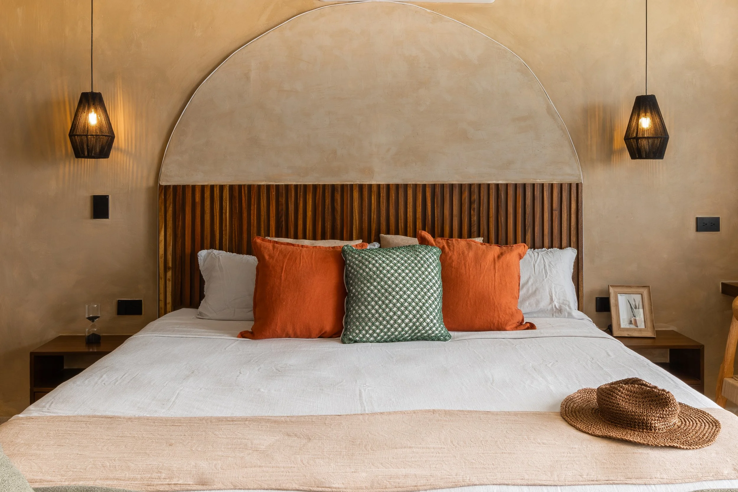 Real estate photography of neatly made bed with orange, green, and white pillows, featuring a wooden headboard with a semi-circular design, at Mahahual.