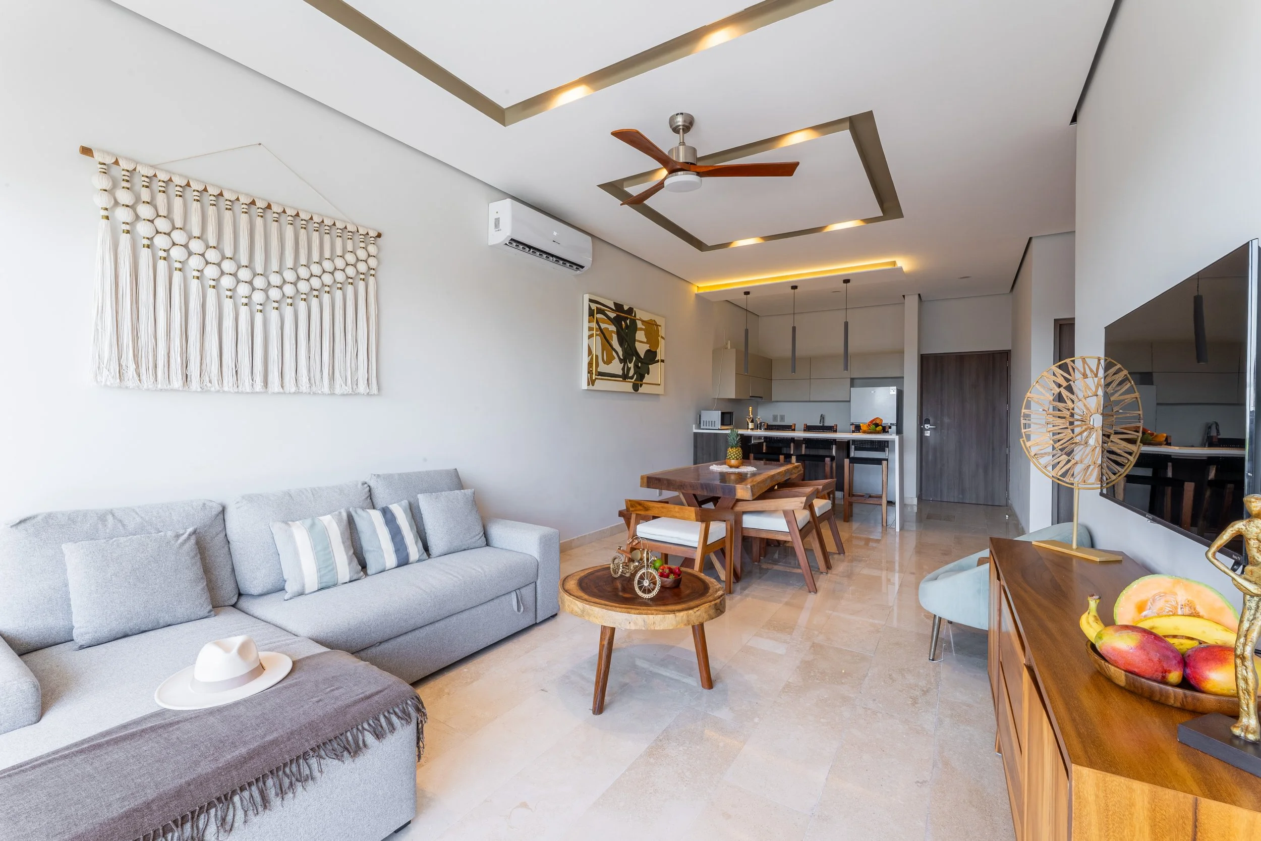 Real estate photography of living room with gray couch, throw blanket, wall art, wooden coffee table with decorative items, dining table with chairs, kitchen in background, ceiling fan, and decorative wall hanging, at Amira Tulum.