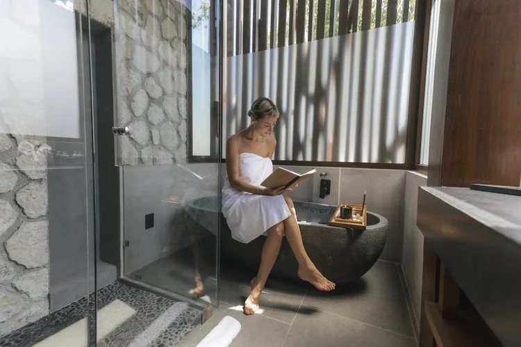 Woman sitting inside a modern bathroom, wrapped in a towel, reading a book or magazine, at Siwa Tulum.