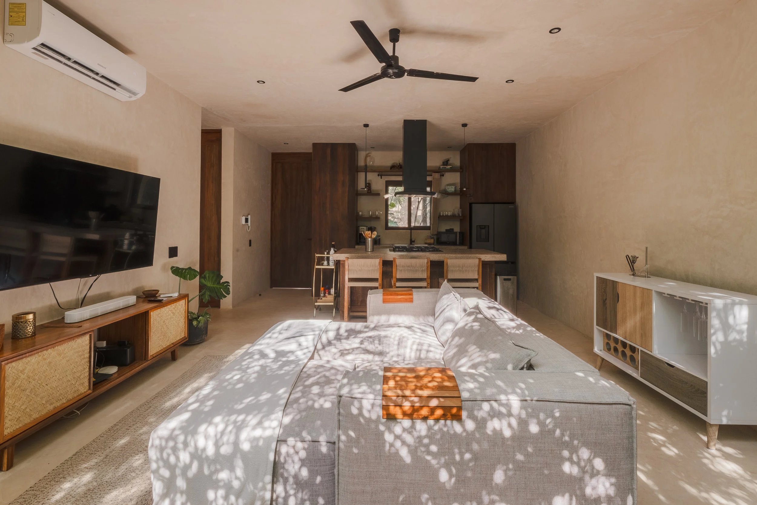 Real estate photography of living room with beige walls, a gray sofa, wooden coffee table, and a TV on a wooden console. Sunlight creates dappled shadows on the furniture, at Tulum.