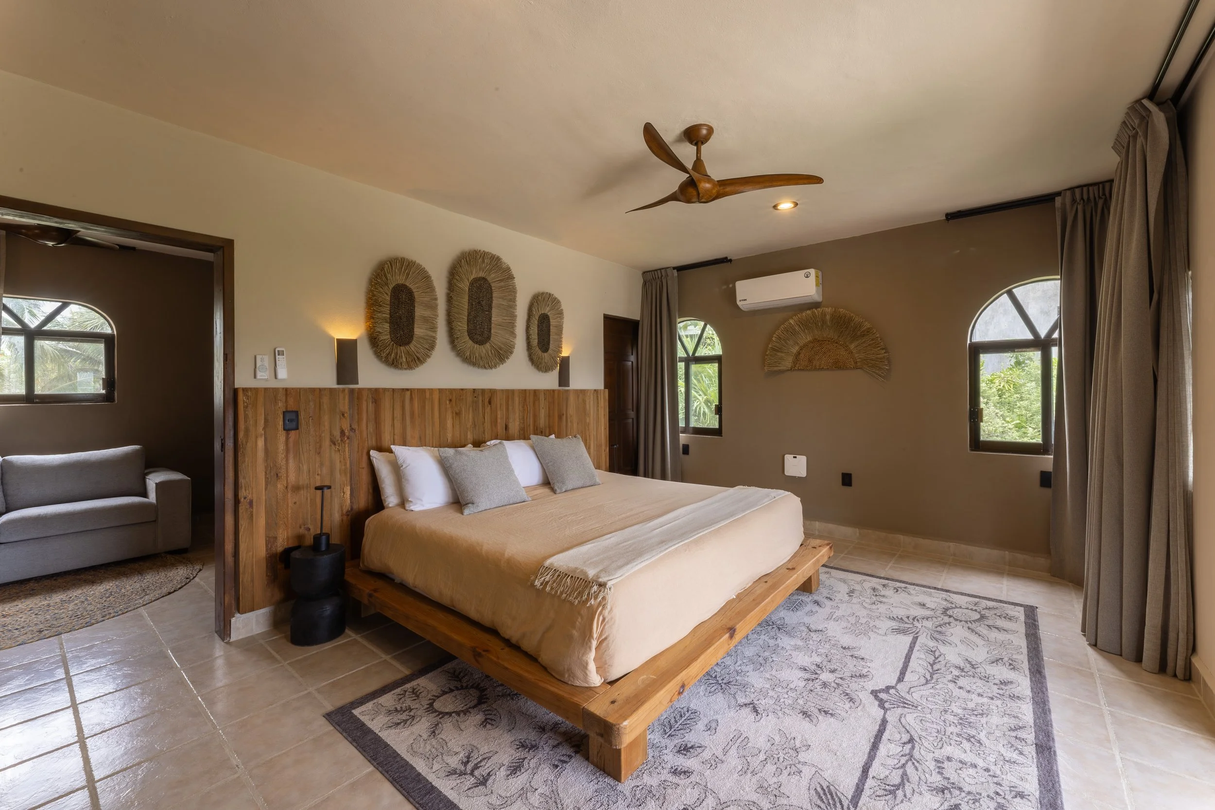 A bedroom with a wooden bed, beige bedding, and pillows, decorated with wall art and an air conditioner, with windows showing greenery outside, at Bespoke Tulum.