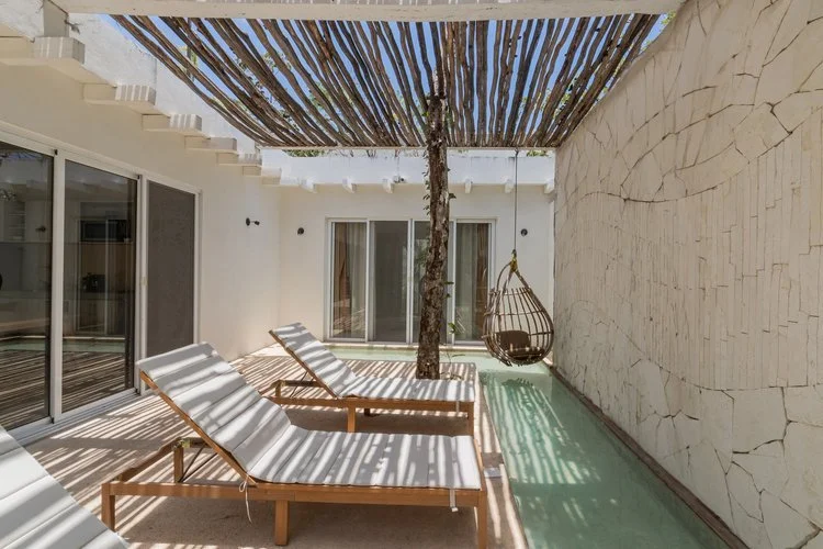 Real estate photography of outdoor patio area with two white lounge chairs, a hanging wicker chair, and a tree, shaded by a wooden pergola, with glass sliding doors and a textured white wall, at Tulum.