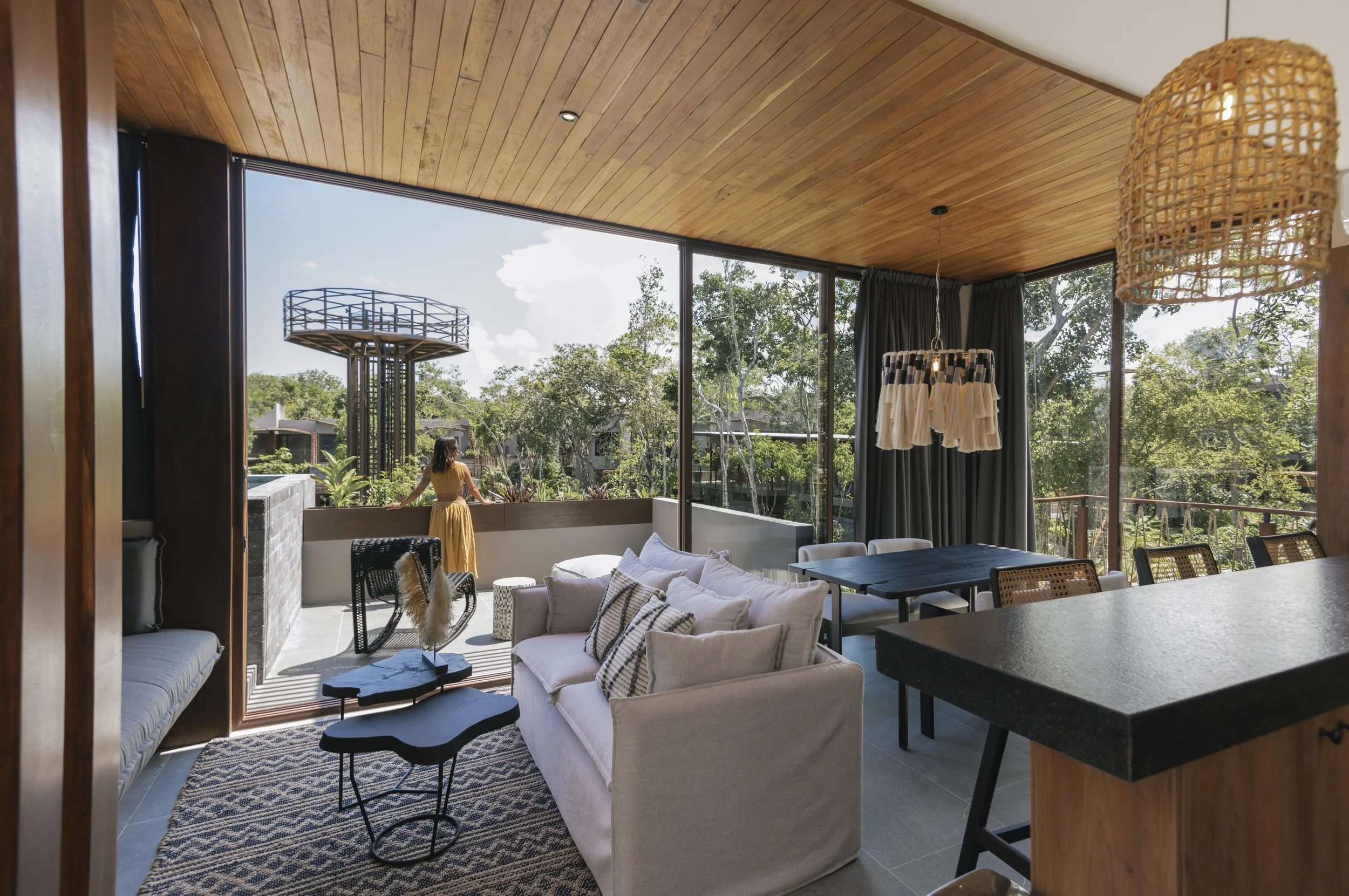 Living room with large glass windows and wooden ceiling, furnished with a white sofa, a black coffee table, a dining table, and chairs, with a woman standing outside on a balcony overlooking trees and a clear sky, at Siwa Tulum.