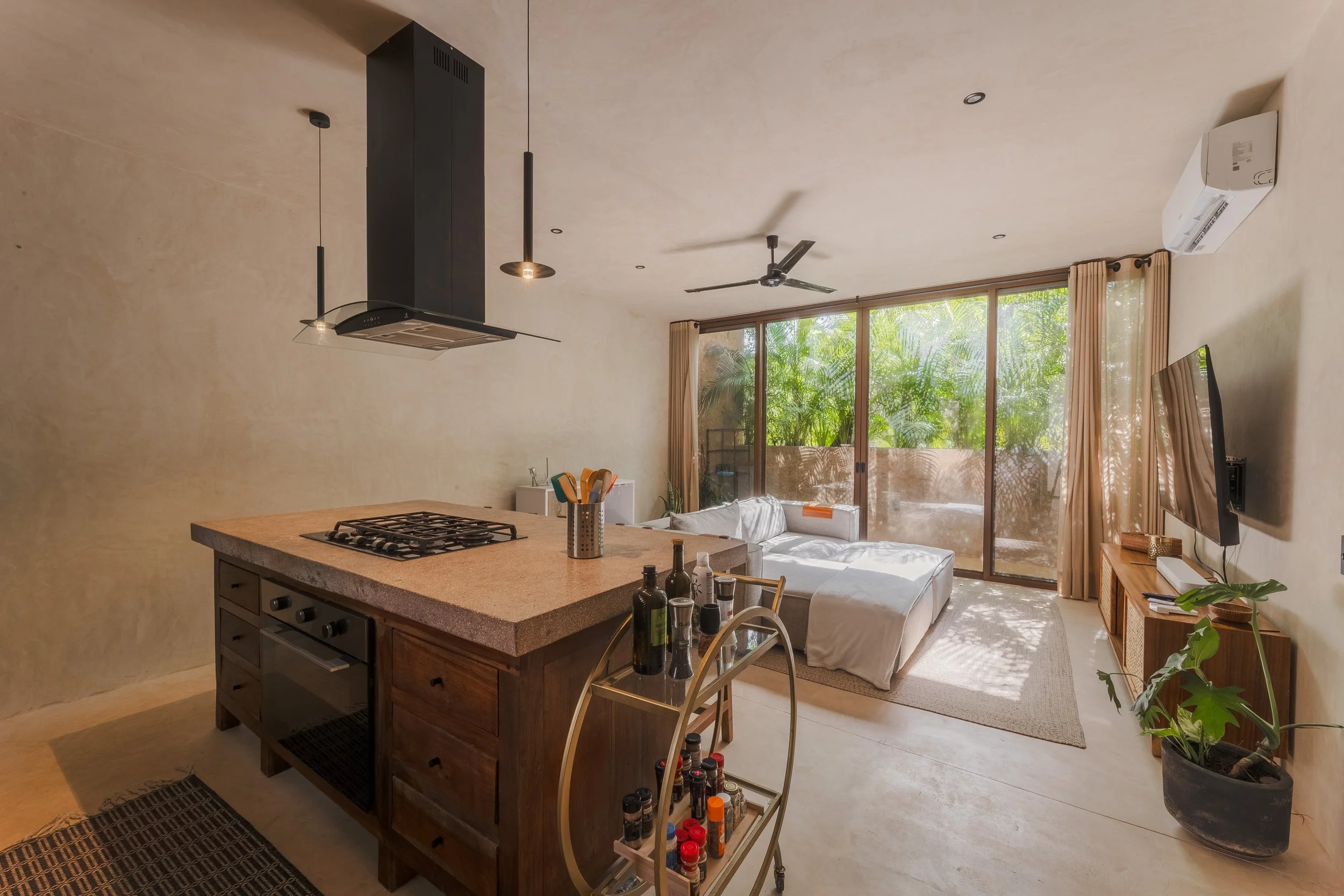 Real estate photography of open-concept living room and kitchen with large windows, beige walls, ceiling fan, mounted air conditioner, and a kitchen island with cooking utensils and bottles, at Tulum.