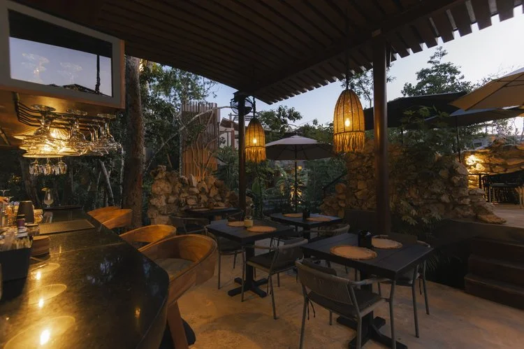 Outdoor restaurant with wooden ceiling, hanging lights, tables with umbrellas, and lush greenery, at Siwa Tulum.
