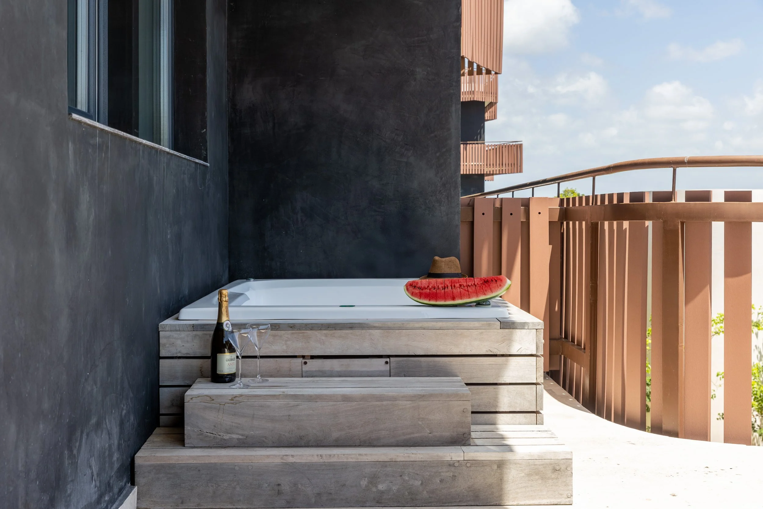 Real estate photography of a balcony with a modern black wall, a wooden bench, a bottle of champagne and two glasses, a large slice of watermelon with a small straw hat on top, and a wooden railing, under a partly cloudy sky, at Amira Tulum.