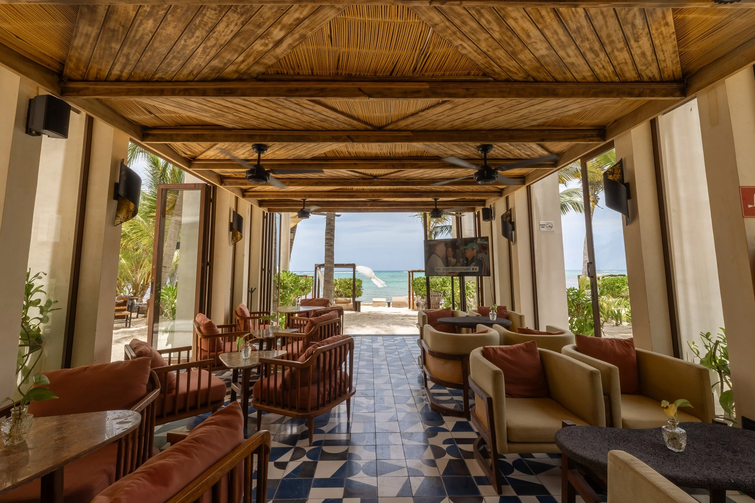 Interior of a beachside restaurant with wooden ceiling, ceiling fans, large open doors, and views of palm trees and the ocean outside, at Bespoke Tulum.