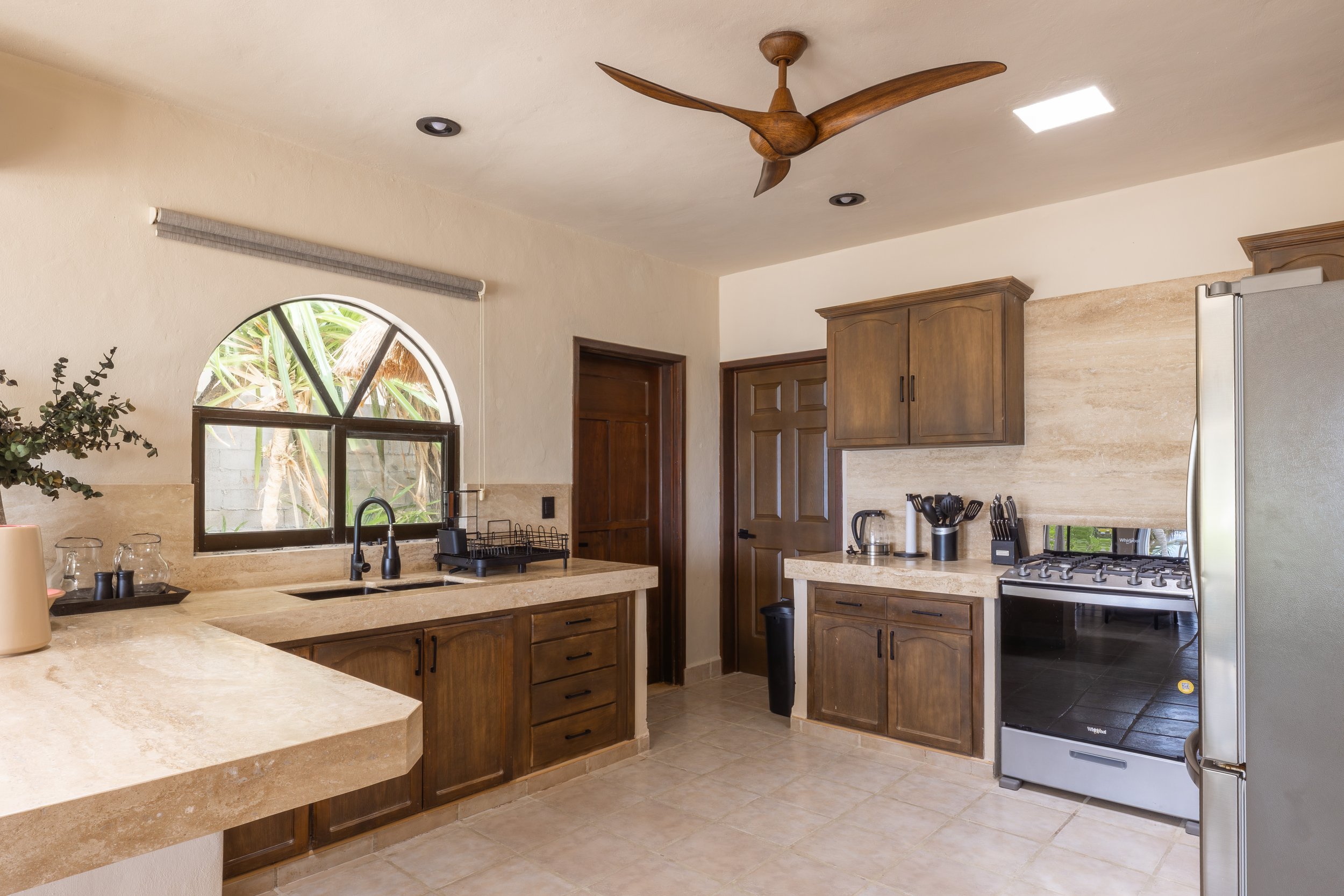 A modern kitchen with wooden cabinets, a beige countertop, a stainless steel refrigerator, and a window with a view of green foliage outside. There is a ceiling fan above and various kitchen utensils on the counter, at Bespoke Tulum.