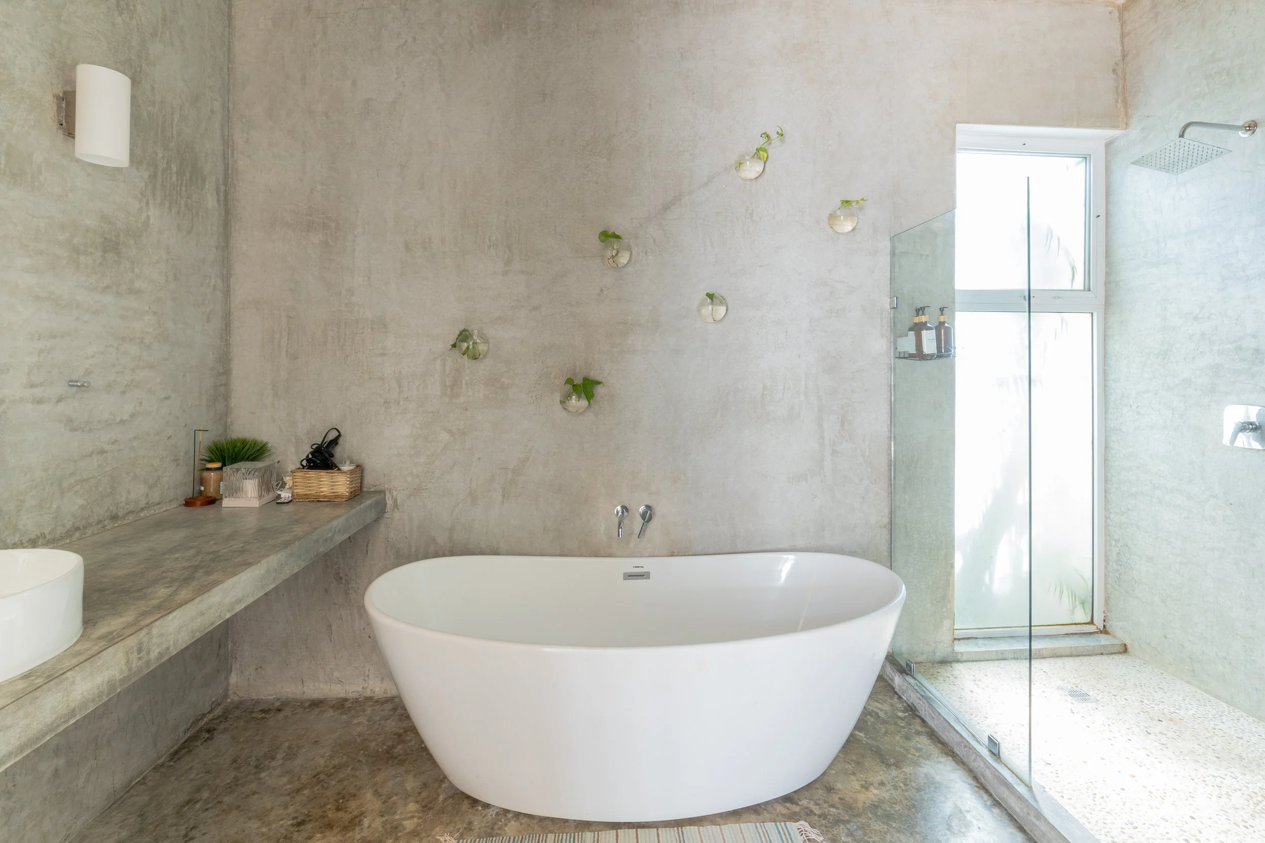 Real estate photography of modern bathroom with a large soaking tub, concrete countertop, and glass-enclosed shower with rainfall showerhead, at Tulum.