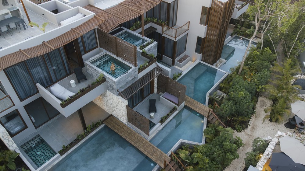 Aerial view of a modern hotel or resort with multiple swimming pools, private balconies with outdoor seating, lush greenery, and pathways, surrounded by trees and tropical plants, at Siwa Tulum.