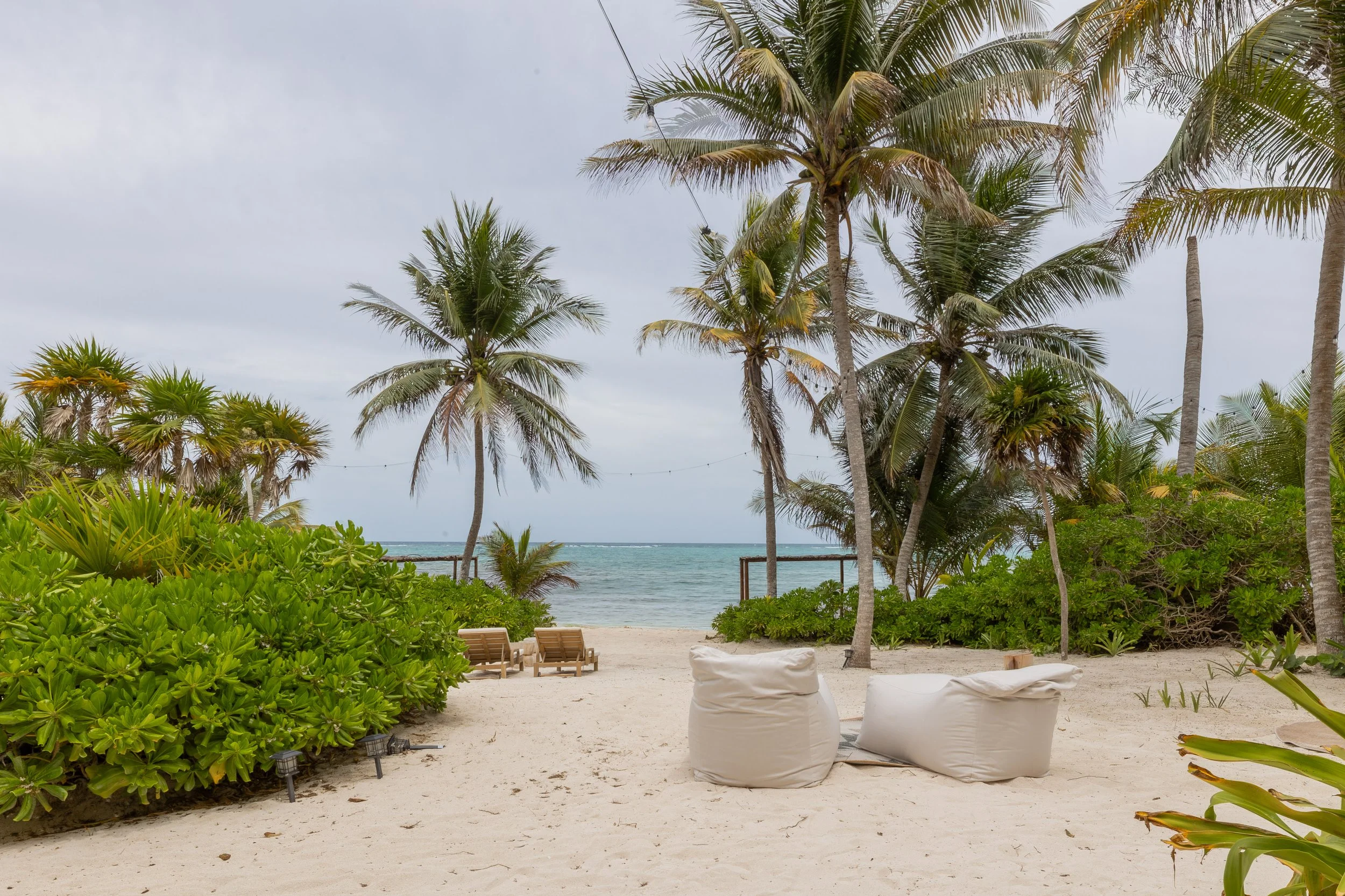 Beachside scene with palm trees, green shrubs, and ocean in the background, featuring outdoor seating with bean bags and lounge chairs under a cloudy sky, at Bespoke Tulum.