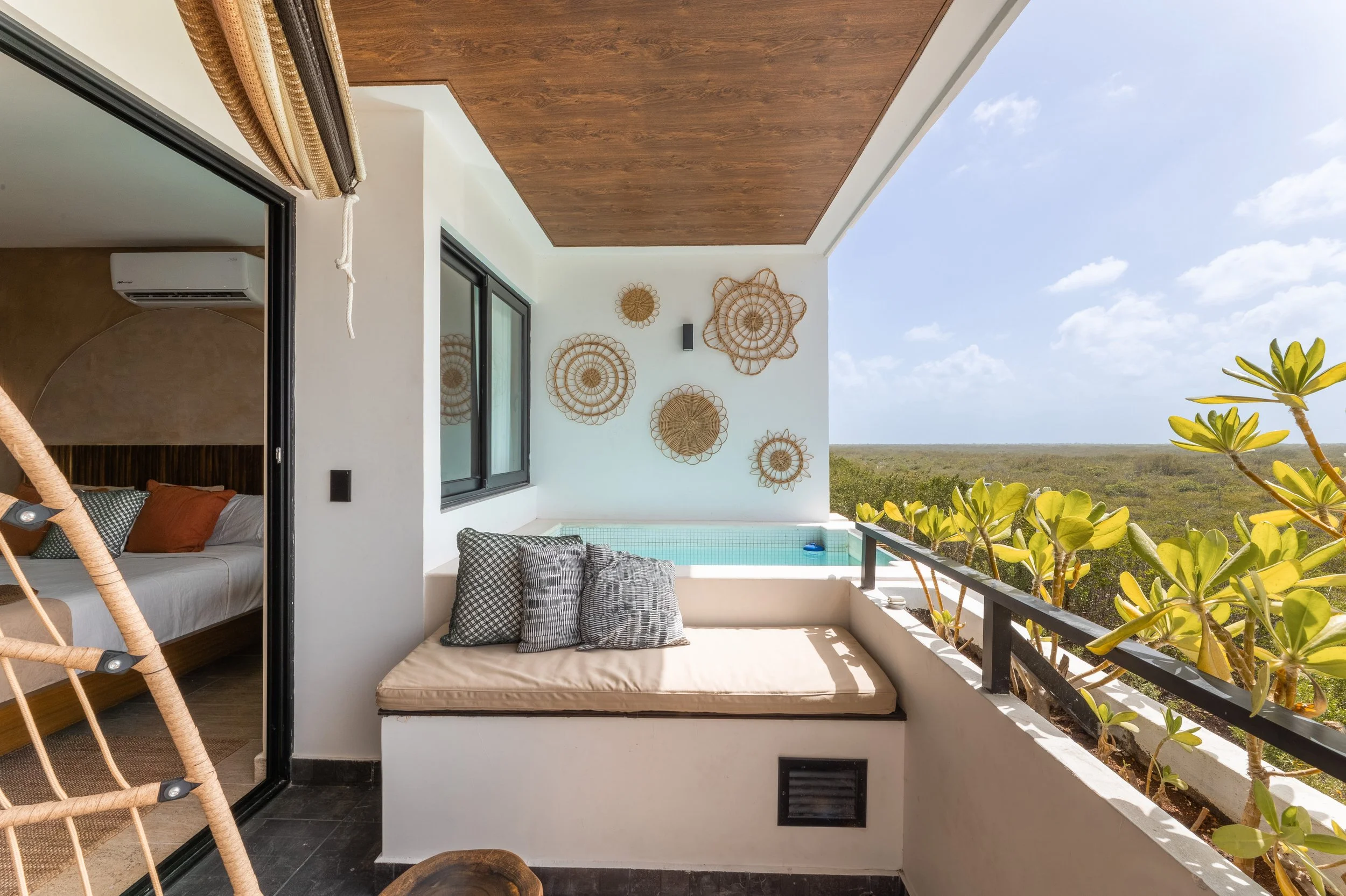 Real estate photography of a modern balcony with a comfortable cushioned seating area, decorative woven wall art, and lush green plants, overlooking a vast natural landscape under a clear blue sky, at Mahahual.
