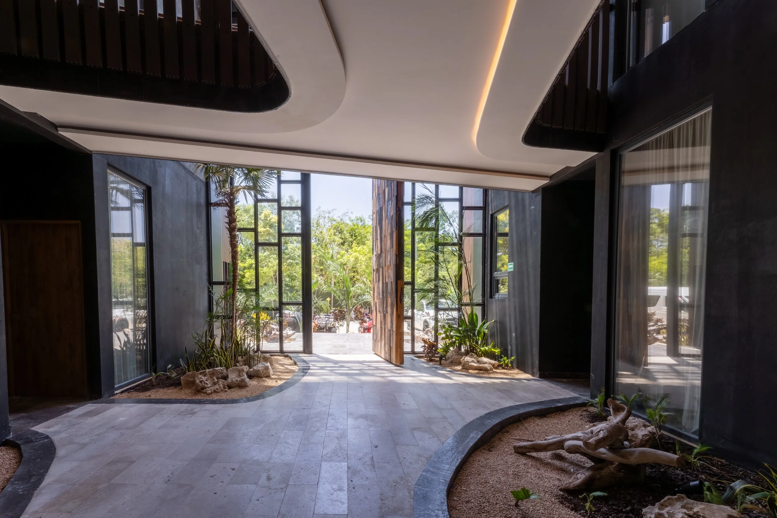 Real estate photography of modern indoor space with large glass windows, potted plants, and a curved pathway leading outside to greenery and trees, at Amira Tulum
