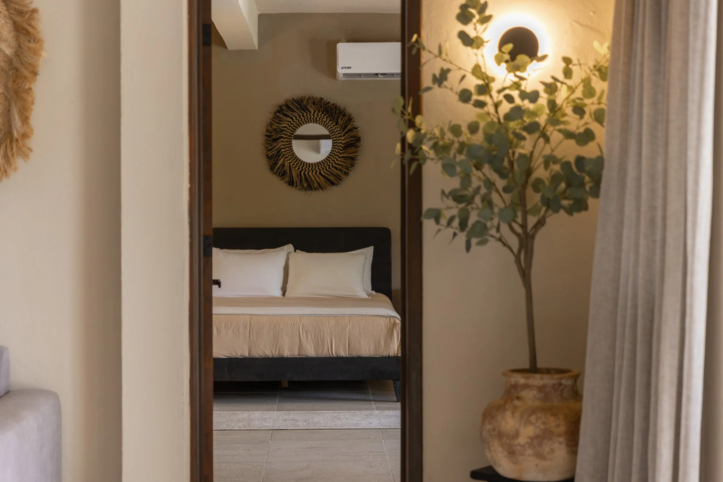 A cozy bedroom with a bed featuring white pillows and bedding, a decorative round mirror with a woven frame on the wall, a potted plant, an air conditioning unit, and warm lighting creating a relaxing ambiance, at Bespoke Tulum.