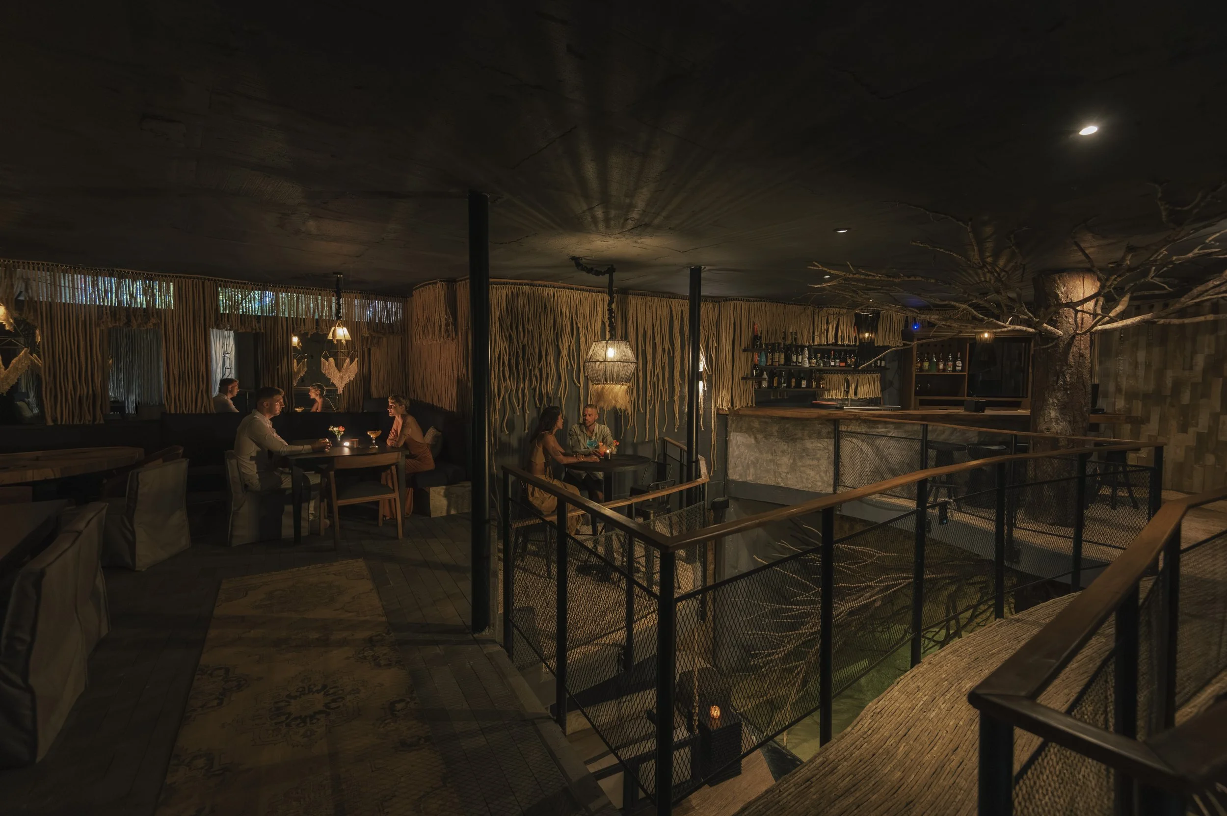Interior of a dimly lit restaurant with wooden decor, pendant lamps, and people sitting at tables, some with drinks, next to a bar area with bottles and a tree branch decoration, at Siwa Tulum.