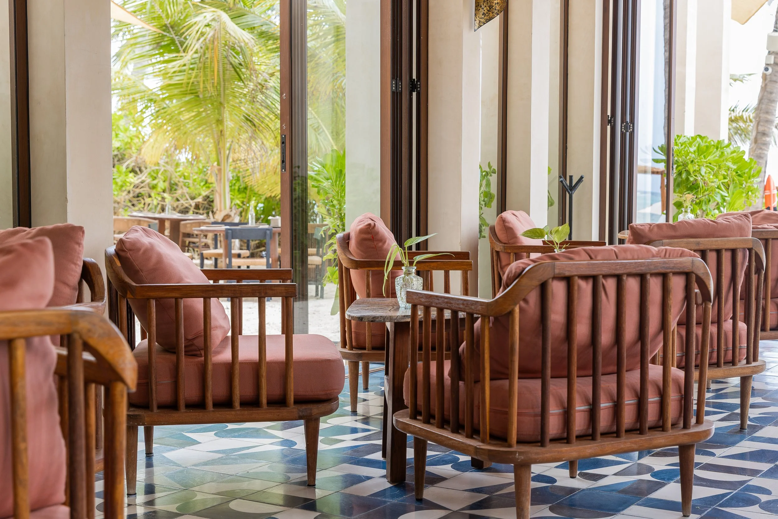 Interior of a cozy lounge with wooden armchairs and pink cushions, large windows revealing tropical greenery outside, and patterned tile floor, at Bespoke Tulum.