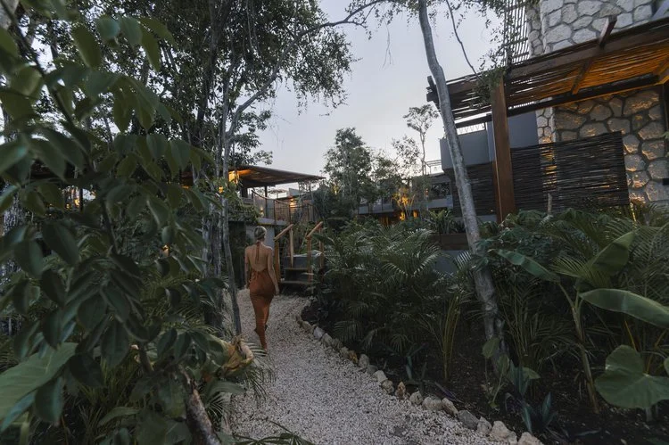 A woman walking along a gravel pathway surrounded by lush green plants, leading towards a modern building with stone walls and wooden accents, in a jungle-like setting during twilight, at Siwa Tulum.