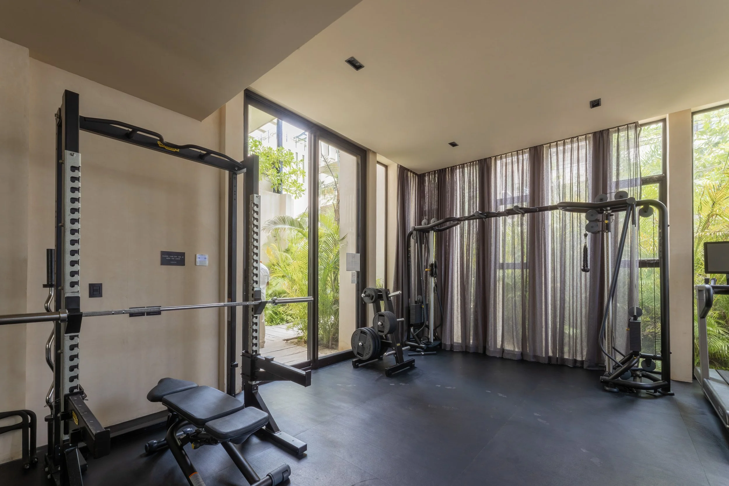 A modern gym with various exercise equipment, large windows with blackout curtains, and greenery visible outside, at Bespoke Tulum.