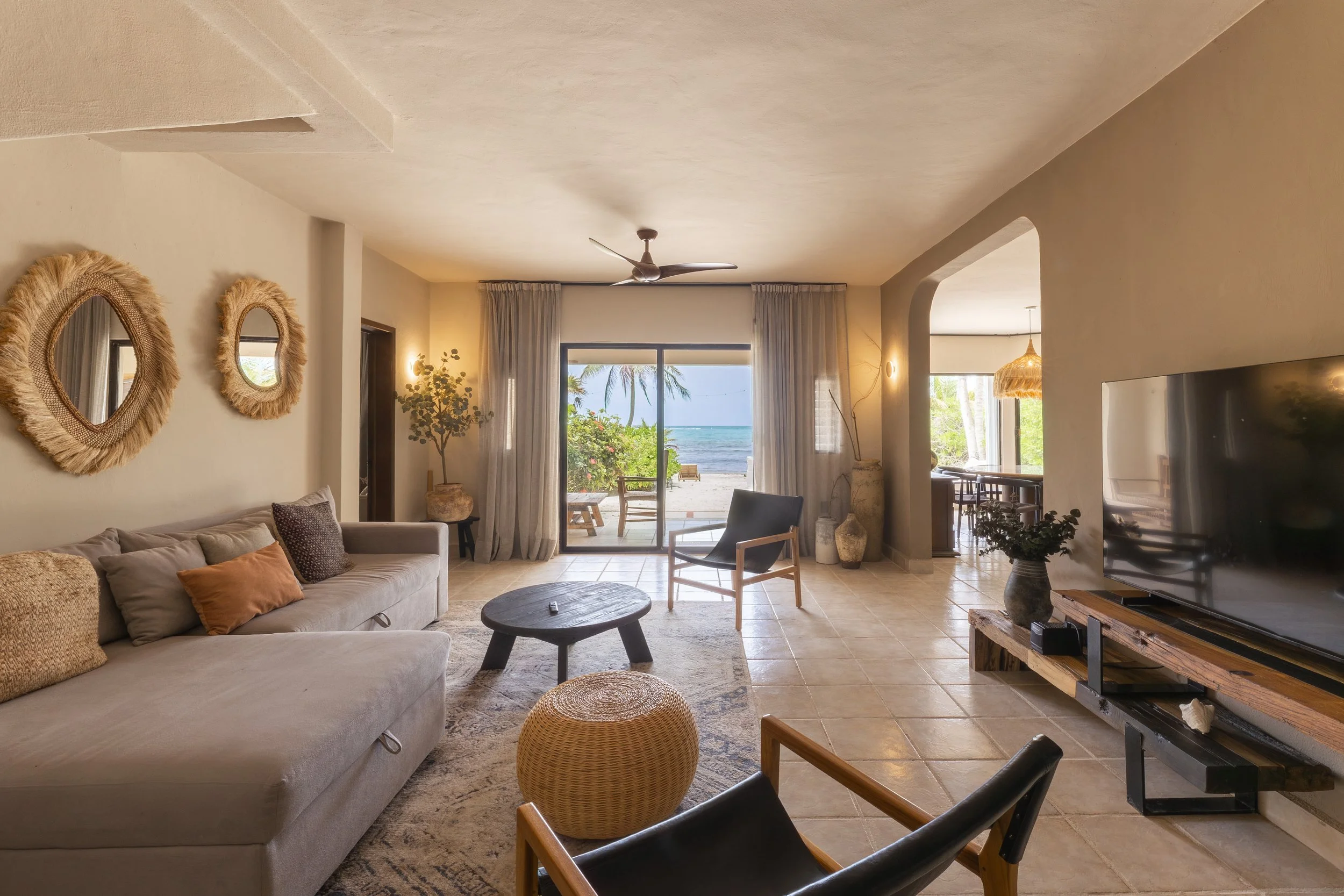 A cozy living room with beige walls and a large flat-screen TV on a wooden stand. . Large sliding glass doors open to a beach view with palm trees and ocean in the background, at Bespoke Tulum.