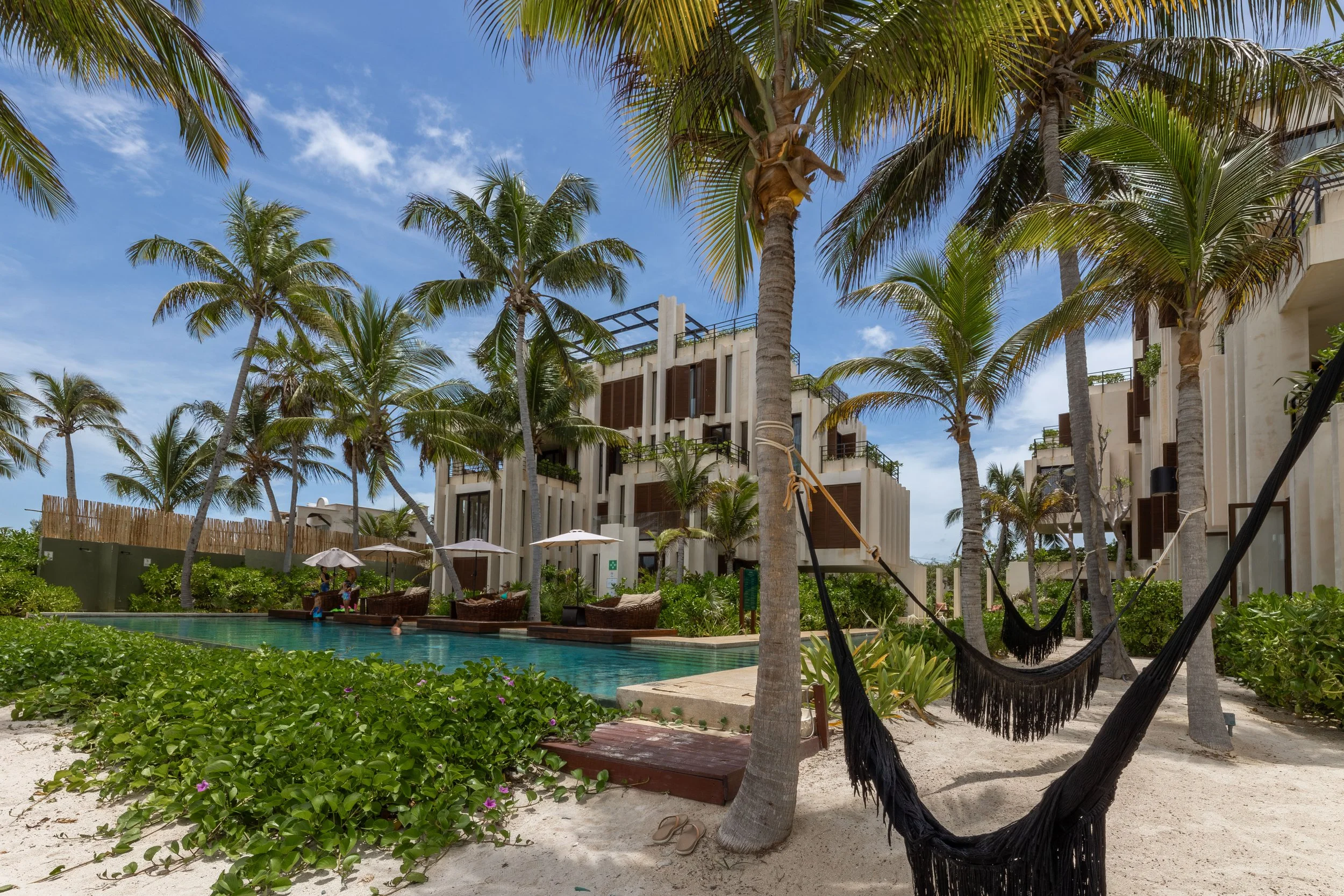A tropical resort scene with palm trees, a swimming pool, and modern white buildings with balconies under a bright blue sky with scattered clouds, at Bespoke Tulum.