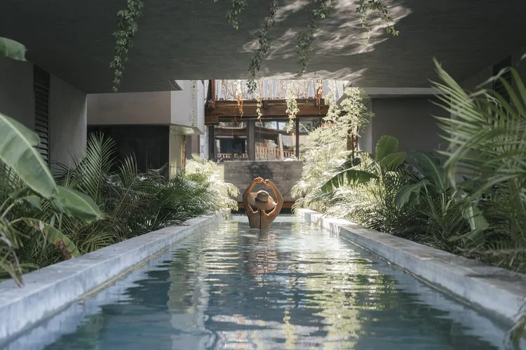 A person relaxing in a narrow indoor pool surrounded by lush tropical plants with an open ceiling and building structure overhead, at Siwa Tulum.
