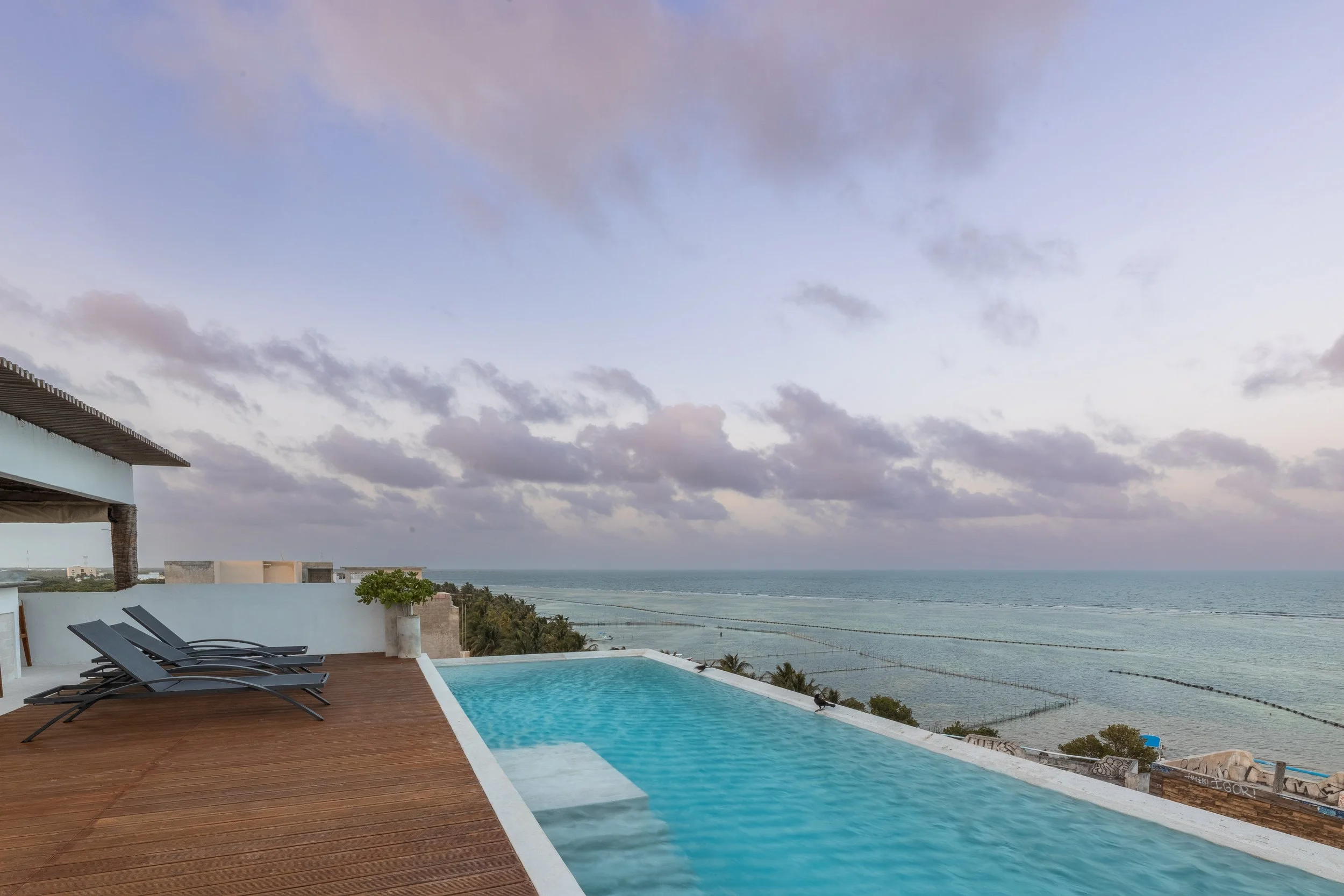 Real estate photography of rooftop infinity pool overlooking the ocean with lounge chairs and a view of the shoreline and cloudy sky, at Mahahual.