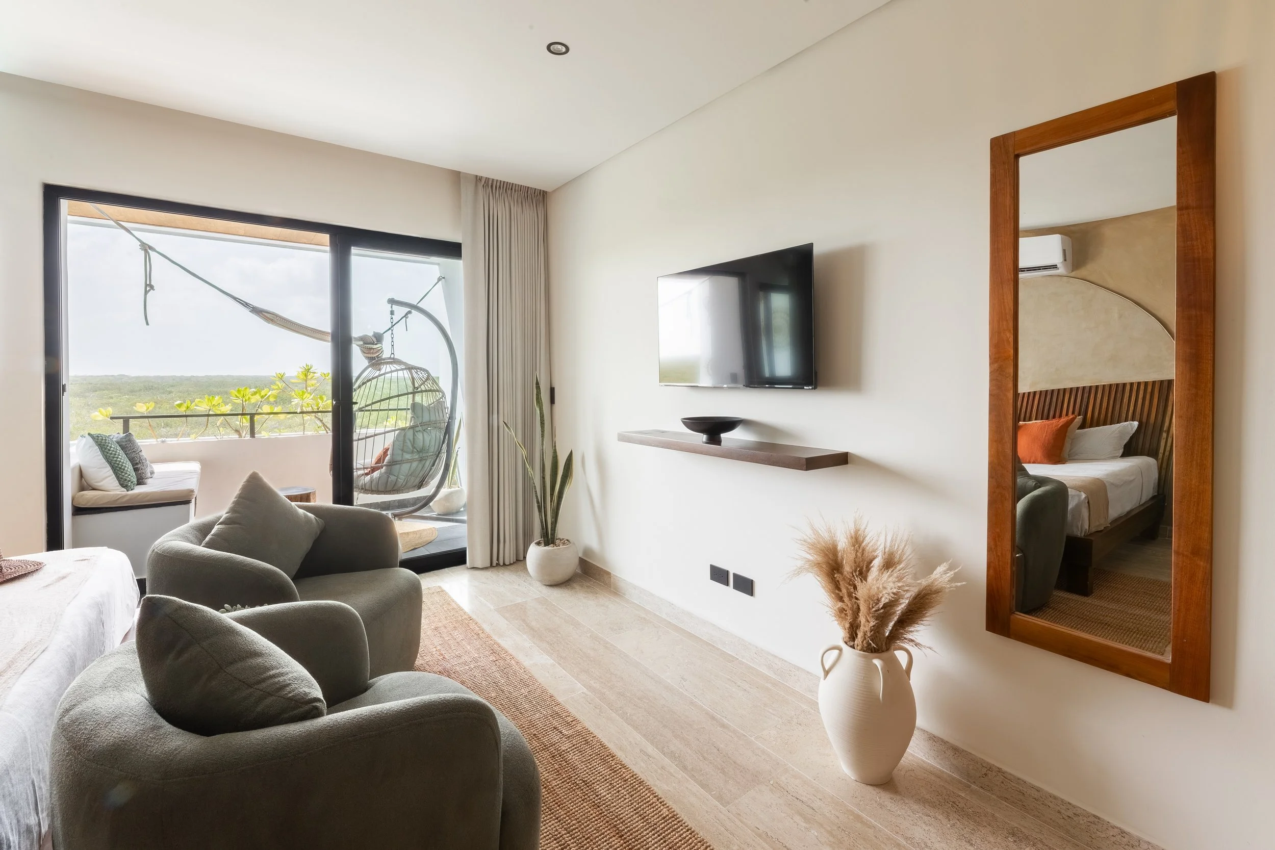 Real estate photography of living room with beige walls, a wall-mounted flat-screen TV, a large mirror in a wooden frame, and sliding glass doors leading to a balcony with outdoor seating and a view of greenery, at Mahahual.