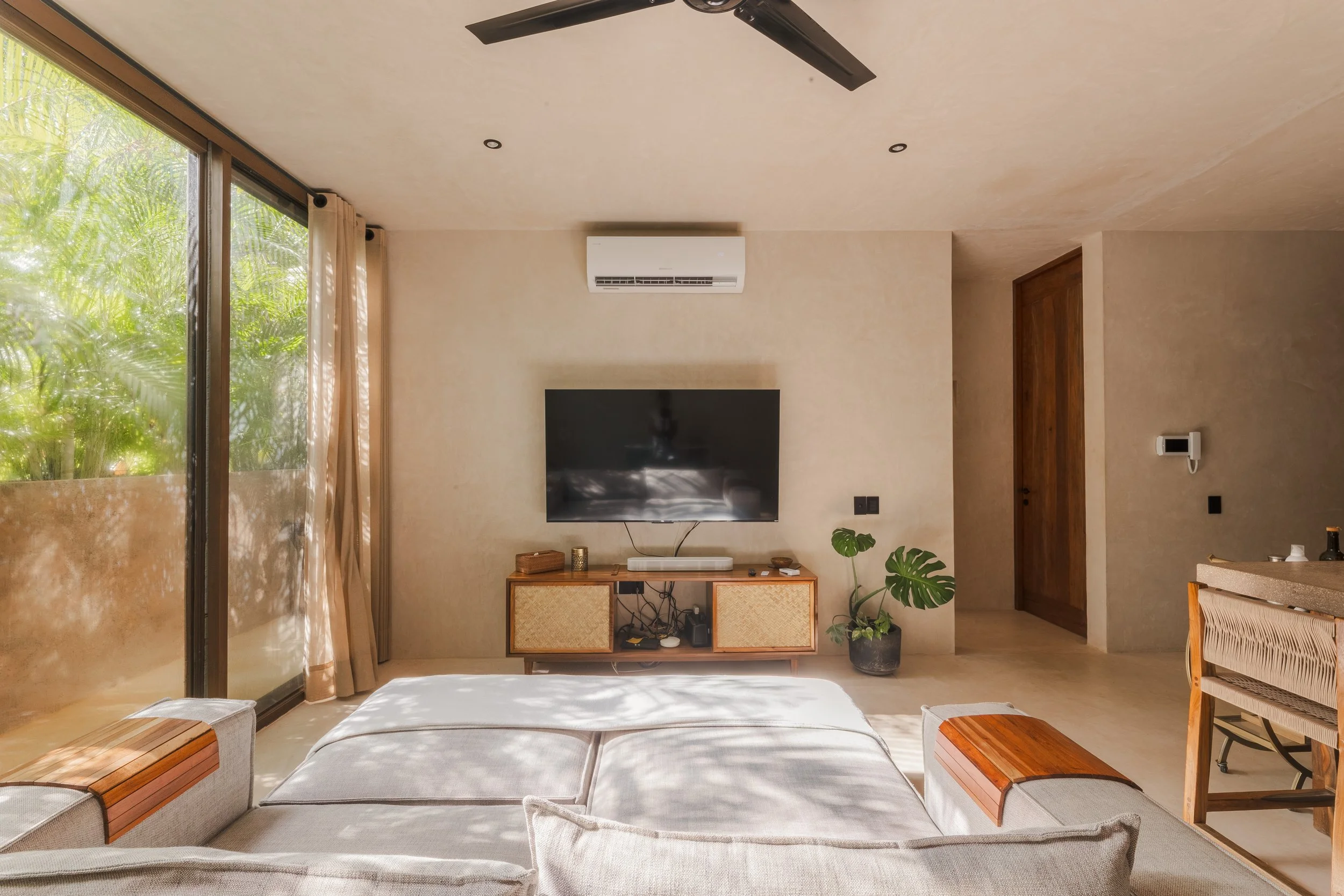 Real estate photography of living room with large sliding glass door showing lush green outdoor plants, beige curtains, a wall-mounted TV, a wooden cabinet, green potted plants, and a ceiling fan, at Tulum