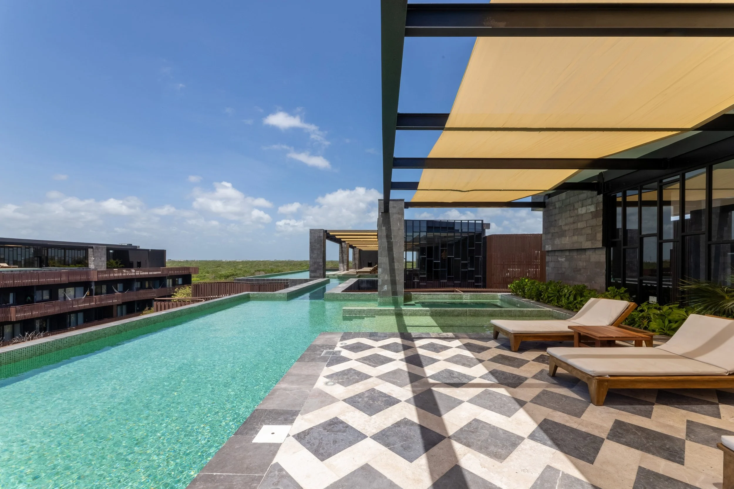 Real estate photography of modern rooftop swimming pool with lounge chairs, a patterned tile deck, and a view of neighboring buildings and blue sky, at Amira Tulum.